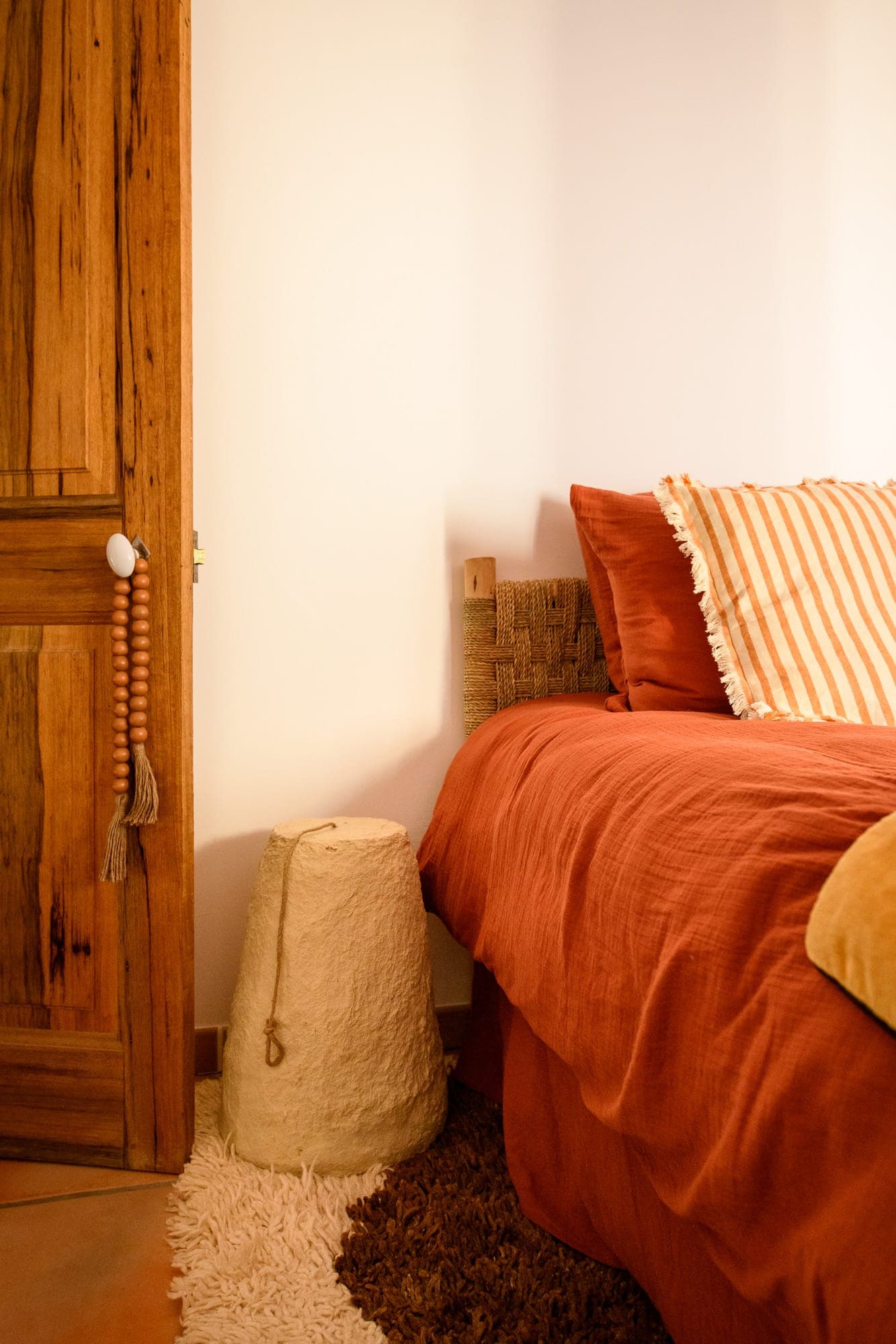 Cosy bedroom with a rustic feel. It features an orange bedspread, striped pillows, a wooden headboard and a wooden door. A textured lamp rests on a soft rug beside the bed, complementing the earthy tones.