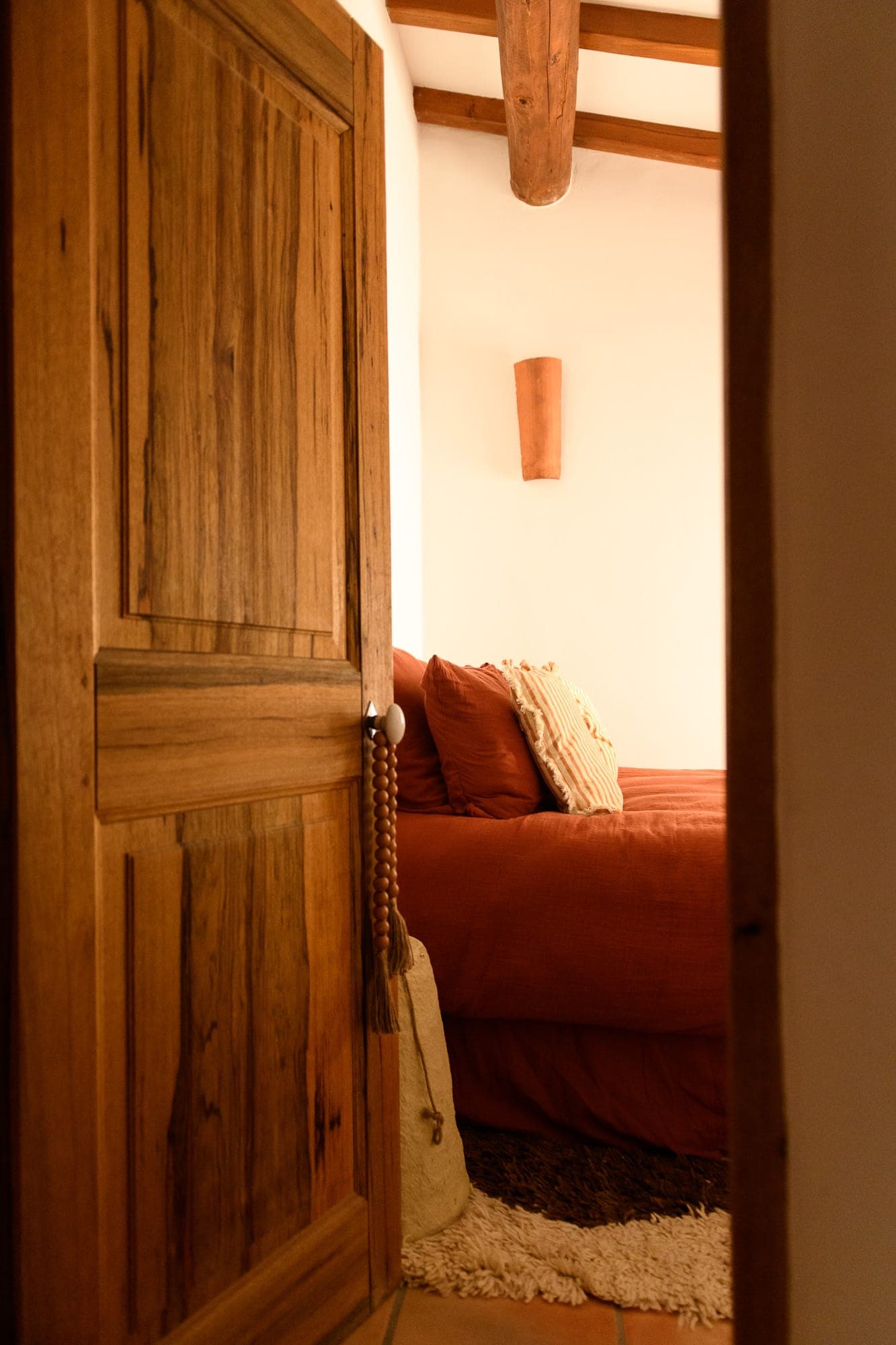 A cozy room seen through a partially open wooden door. The room features a bed with rust-colored bedding, a striped pillow, a soft rug and warm lighting, creating a welcoming, rustic atmosphere.