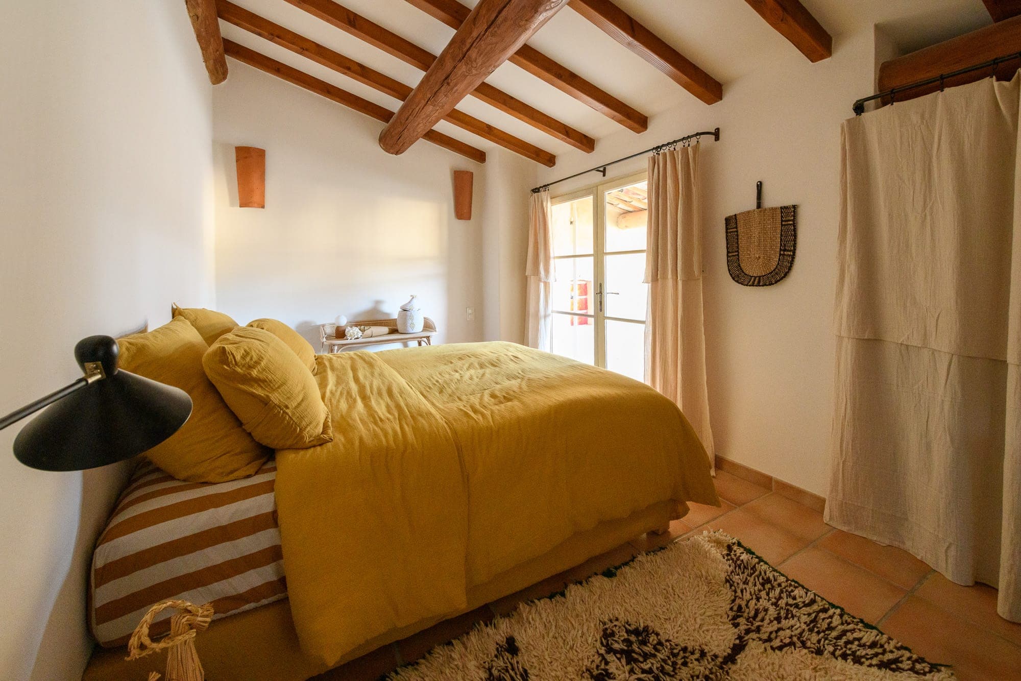 A comfortable room with a wood-beamed ceiling and a bed dressed in yellow sheets. Natural light streams in through a glass door framed by light curtains. A small side table holds decorative objects and a woven wall hanging adds texture.