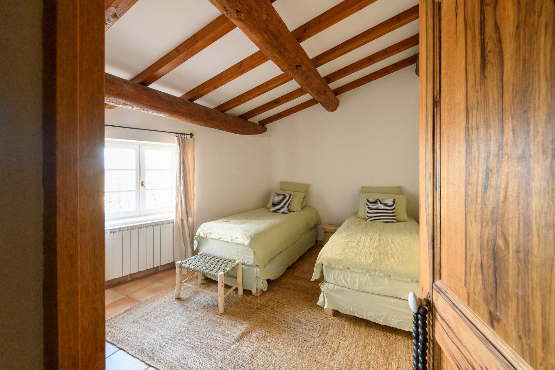 A comfortable room with two single beds with light green bedding, wooden beams on the ceiling, a window with transparent curtains, a wooden door and a woven carpet on the floor. A small bench sits at the foot of one bed.