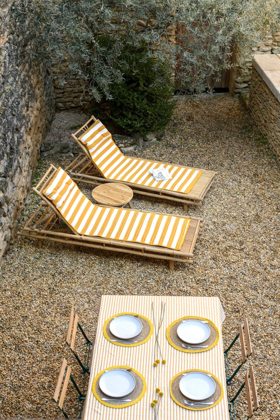 A warm outdoor space with two yellow-and-white striped deckchairs next to a stone wall and a small round table. In the foreground, a dining table is set with four plates and placemats, in harmony with the colors of the deckchairs. The whole is surrounded by pebbles and greenery.