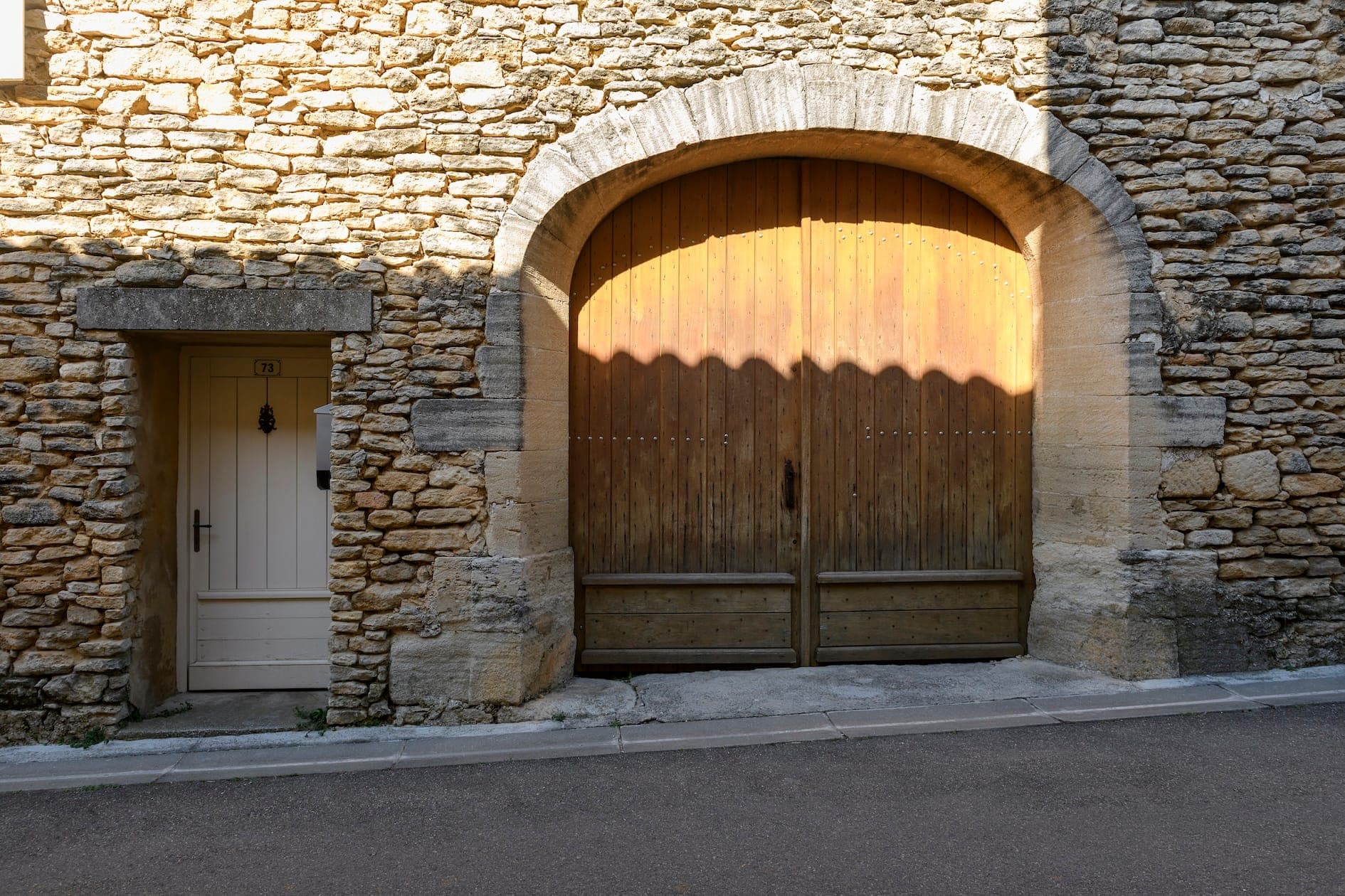 A large, arched wooden door is set into a rustic stone wall, next to a smaller white door. Sunlight casts a shadow on the wooden door, highlighting its textured surface. A cobbled street runs in front of the building.