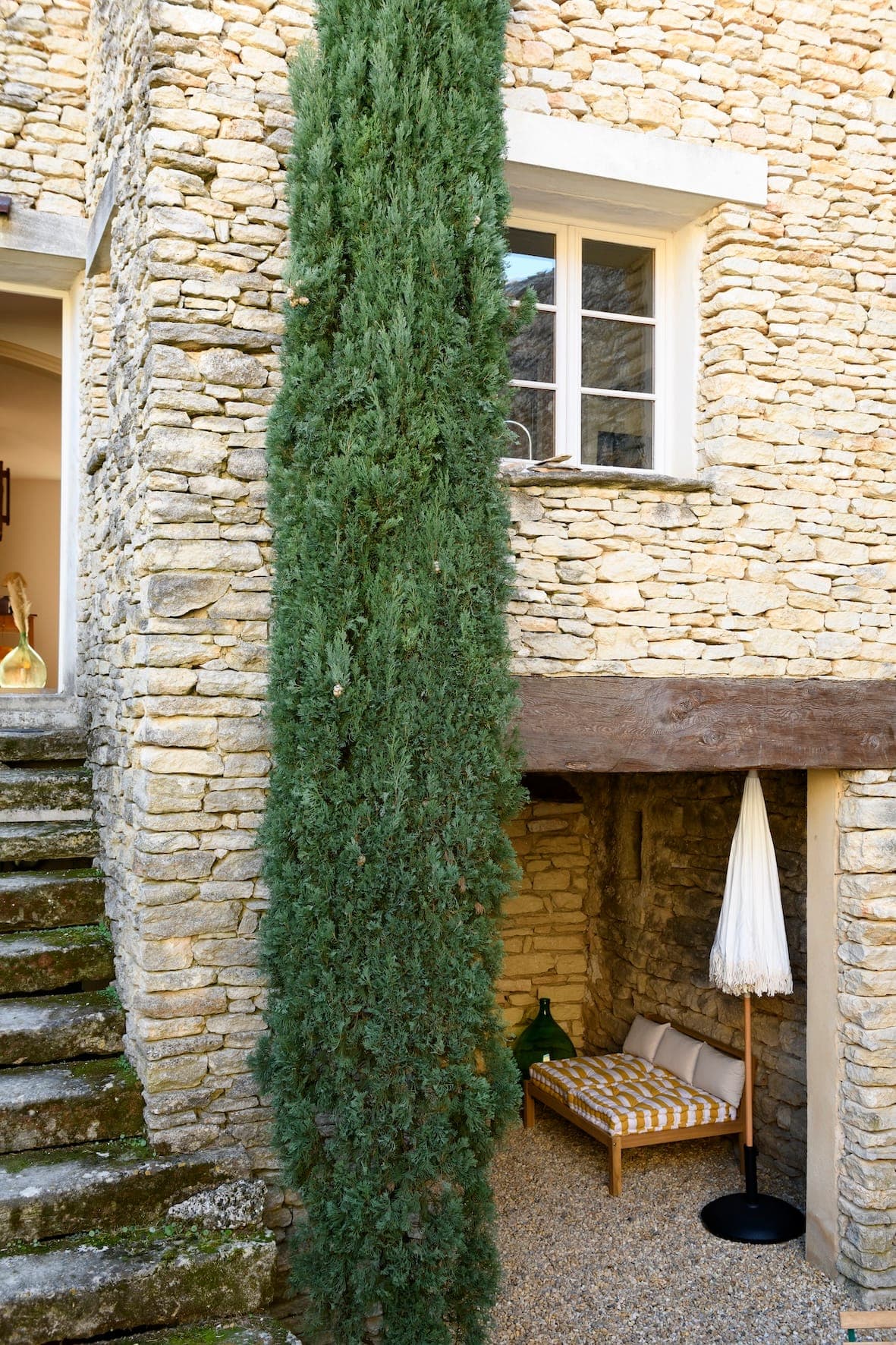 A tall, narrow evergreen tree stands next to a stone building. Beneath a window, a small patio features an upholstered bench with a striped cover and a closed white parasol. Stone steps lead to an entrance on the left.