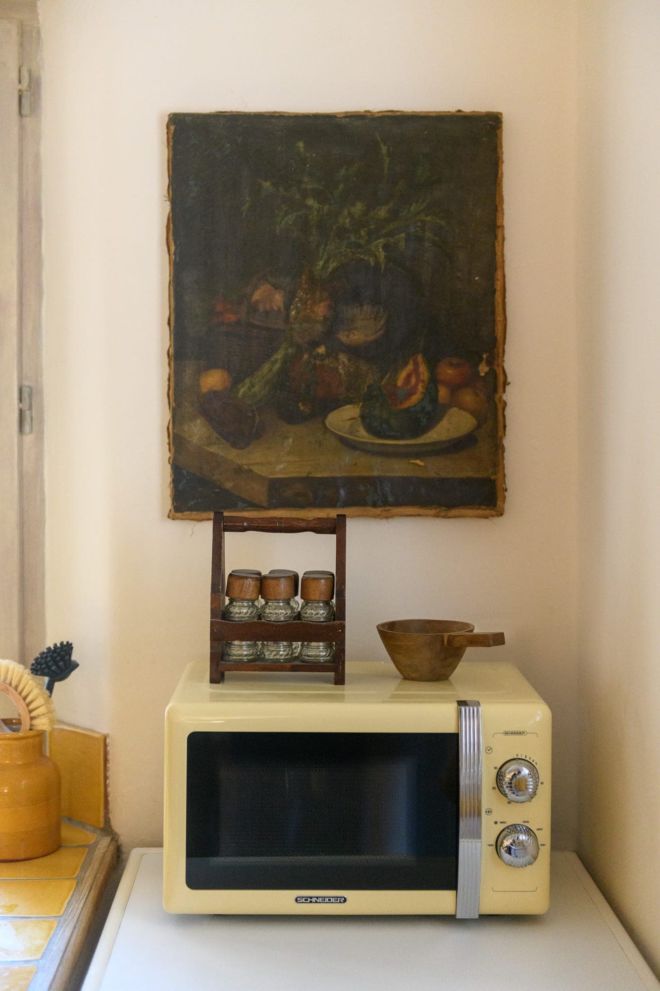 A vintage cream-colored microwave sits on a countertop with a wooden spice rack and wooden bowl on top. Above, a still life in dark tones depicting fruit and a vase hangs on the wall. A yellow ceramic teapot stands in the corner.