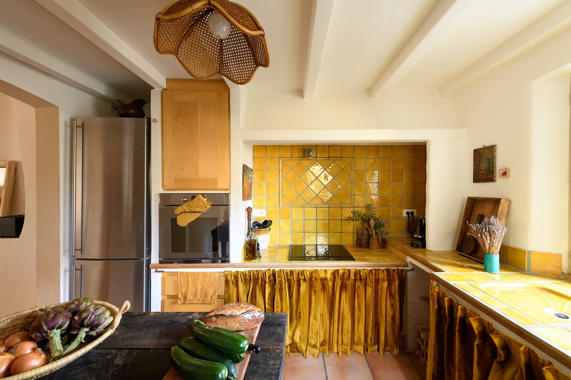 Warm, rustic-style kitchen with backsplash and yellow-tiled countertops. A stainless steel refrigerator sits next to wood cabinets. The island is stocked with fresh vegetables and a wicker light fixture hangs above. An artichoke basket is in the foreground.