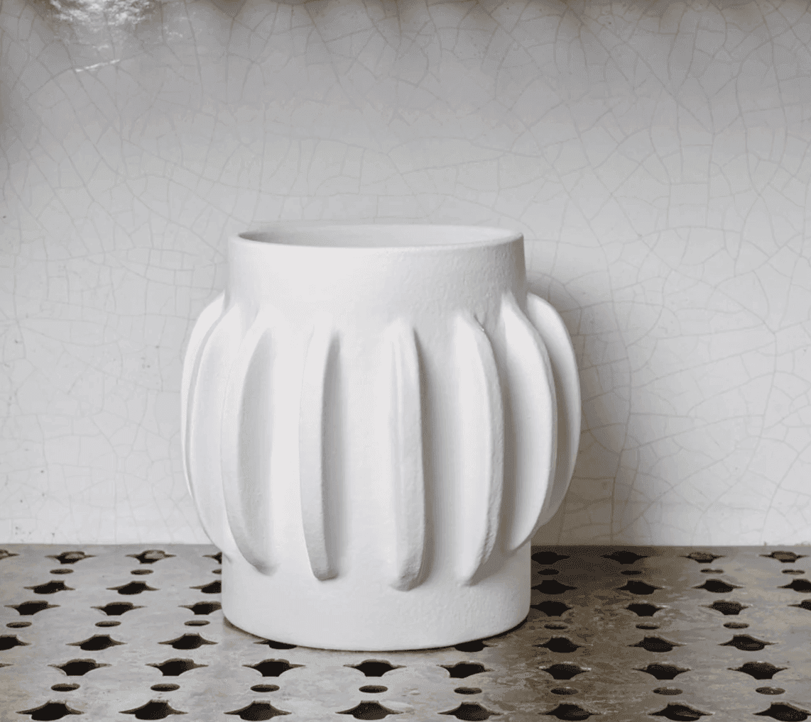 Un vase en céramique blanche avec des crêtes verticales repose sur une surface métallique texturée et perforée contre un mur blanc fissuré.