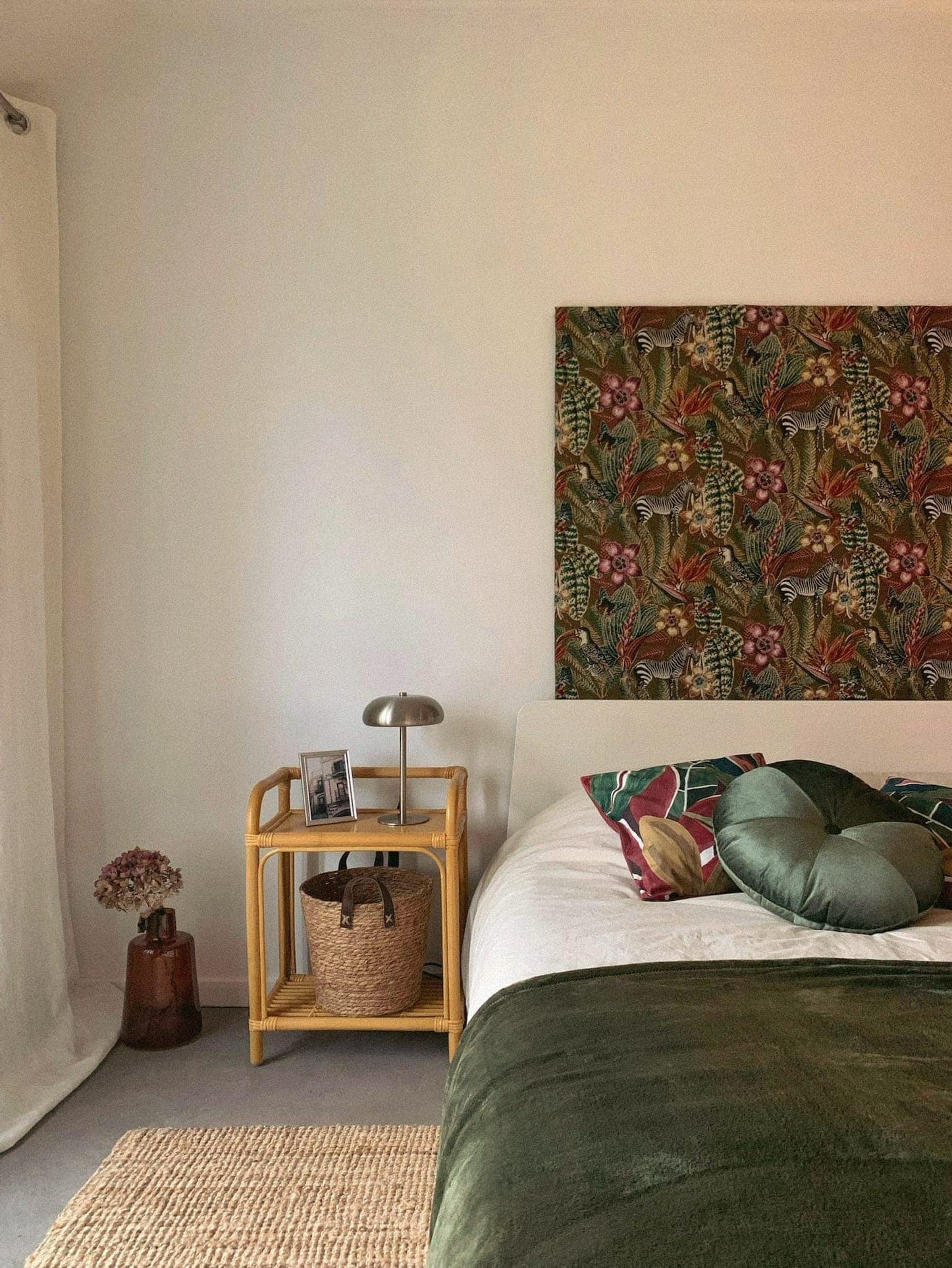 Une chambre confortable comprend un lit avec des oreillers verts et fleuris et une décoration murale à thème tropical au-dessus. Une petite table d'appoint en bois accueille une lampe, une photo encadrée et un panier. Un rideau simple et des fleurs séchées rehaussent l'ambiance sereine.