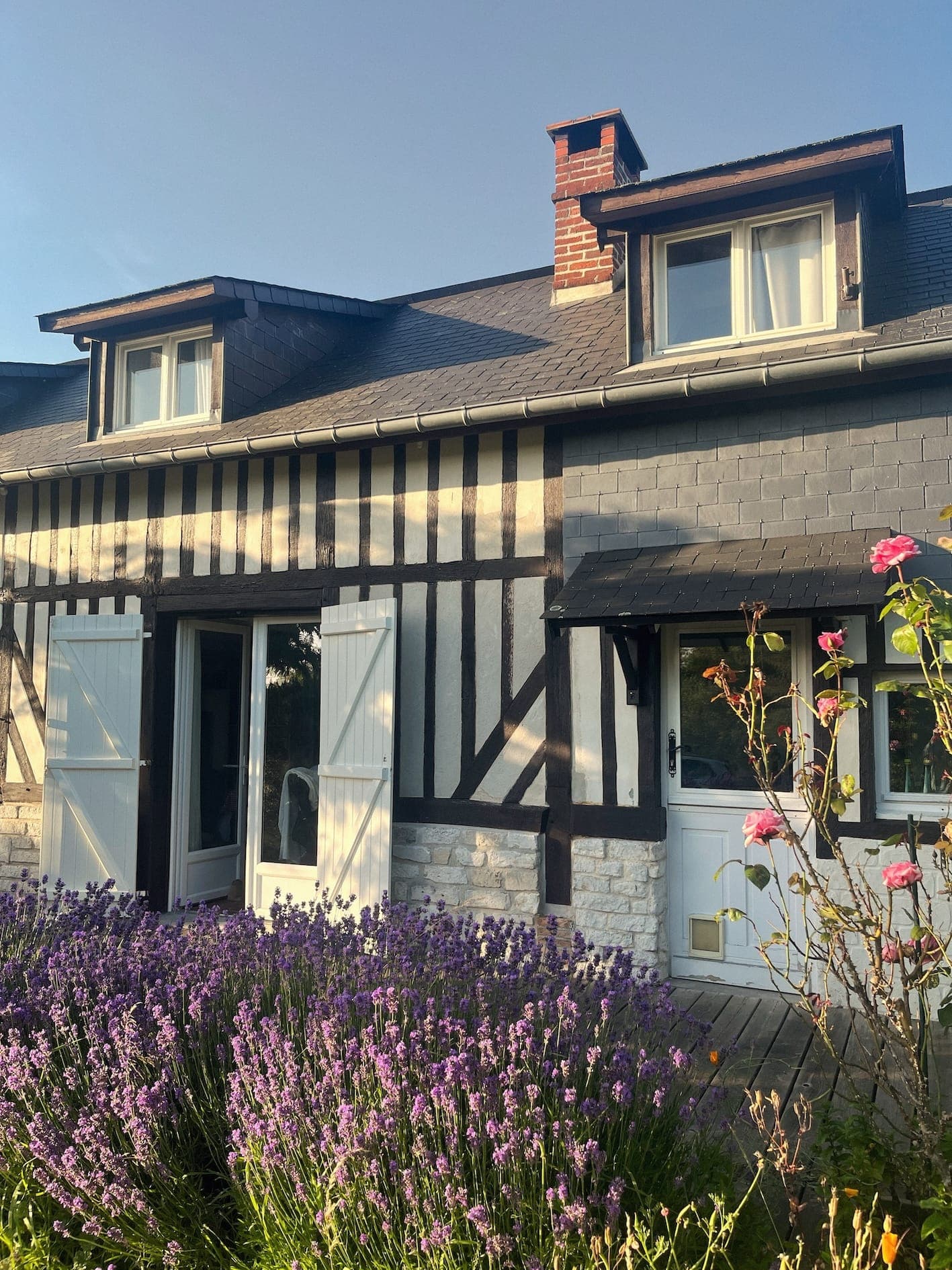 Une charmante maison à colombages aux volets blancs est entourée de lavande en fleurs et de roses roses. Le ciel est clair et la lumière du soleil projette de douces ombres sur le bâtiment.