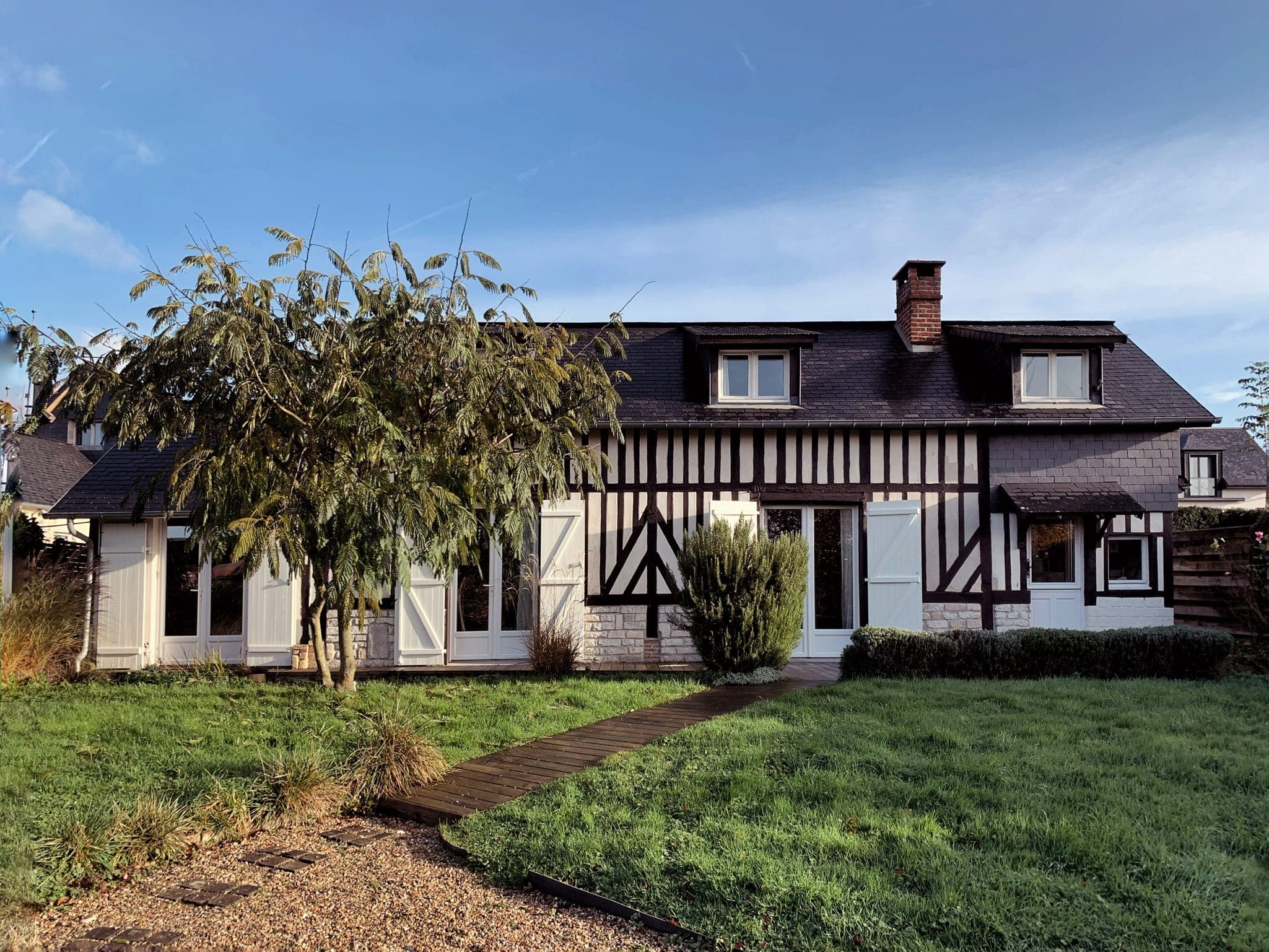 A charming half-timbered house with white and dark brown accents is set against a blue sky. It features several windows, a chimney, and a lush green lawn with a small tree and a gravel pathway leading to the entrance.