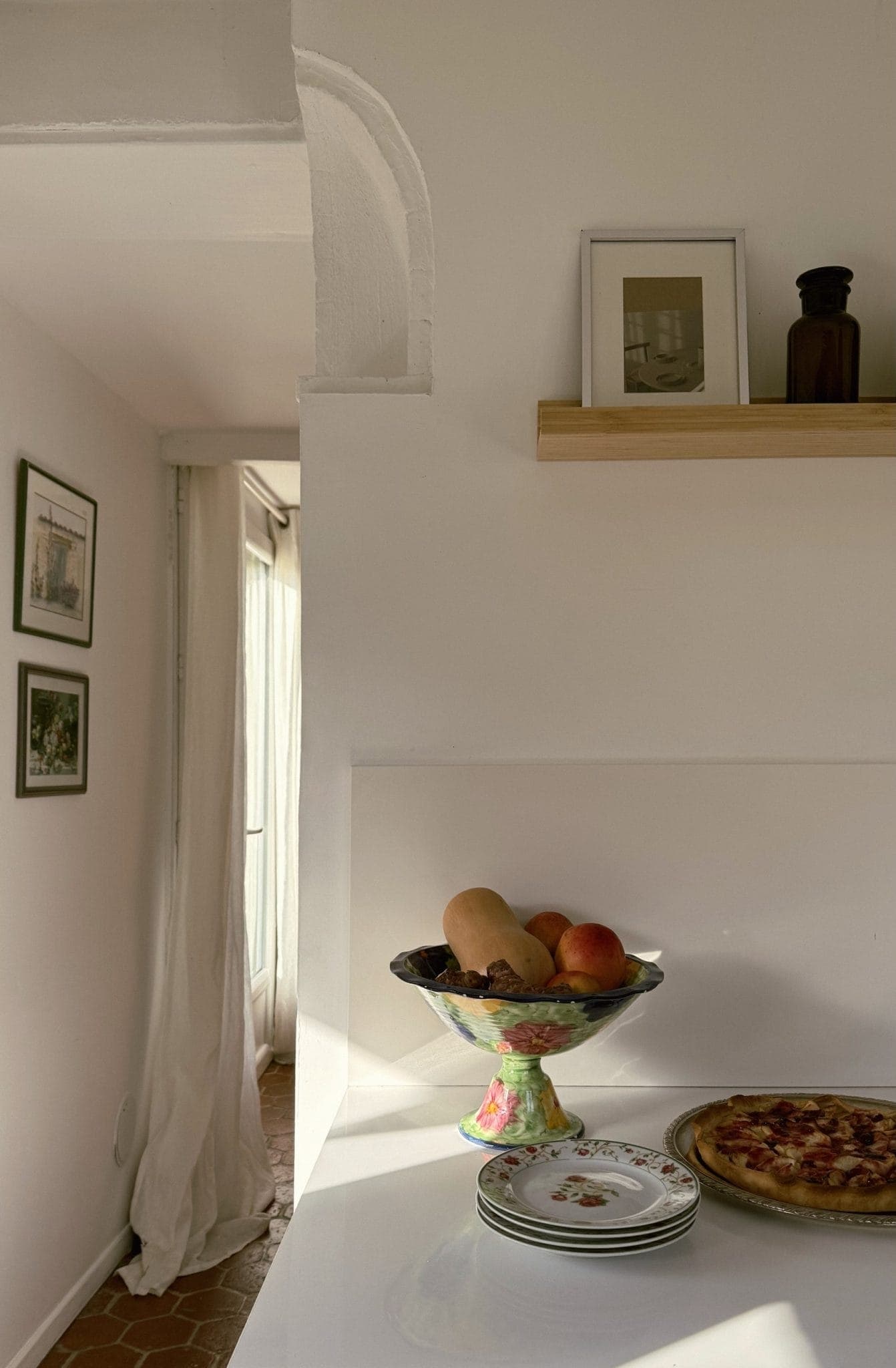 Une cuisine minimaliste aux murs blancs et une étagère sur laquelle sont exposées une photo encadrée et une bouteille. Un plan de travail abrite un bol décoratif contenant des légumes et des fruits, des assiettes à motifs floraux et une pizza. La lumière du soleil entre par une fenêtre voisine dotée de rideaux drapés.