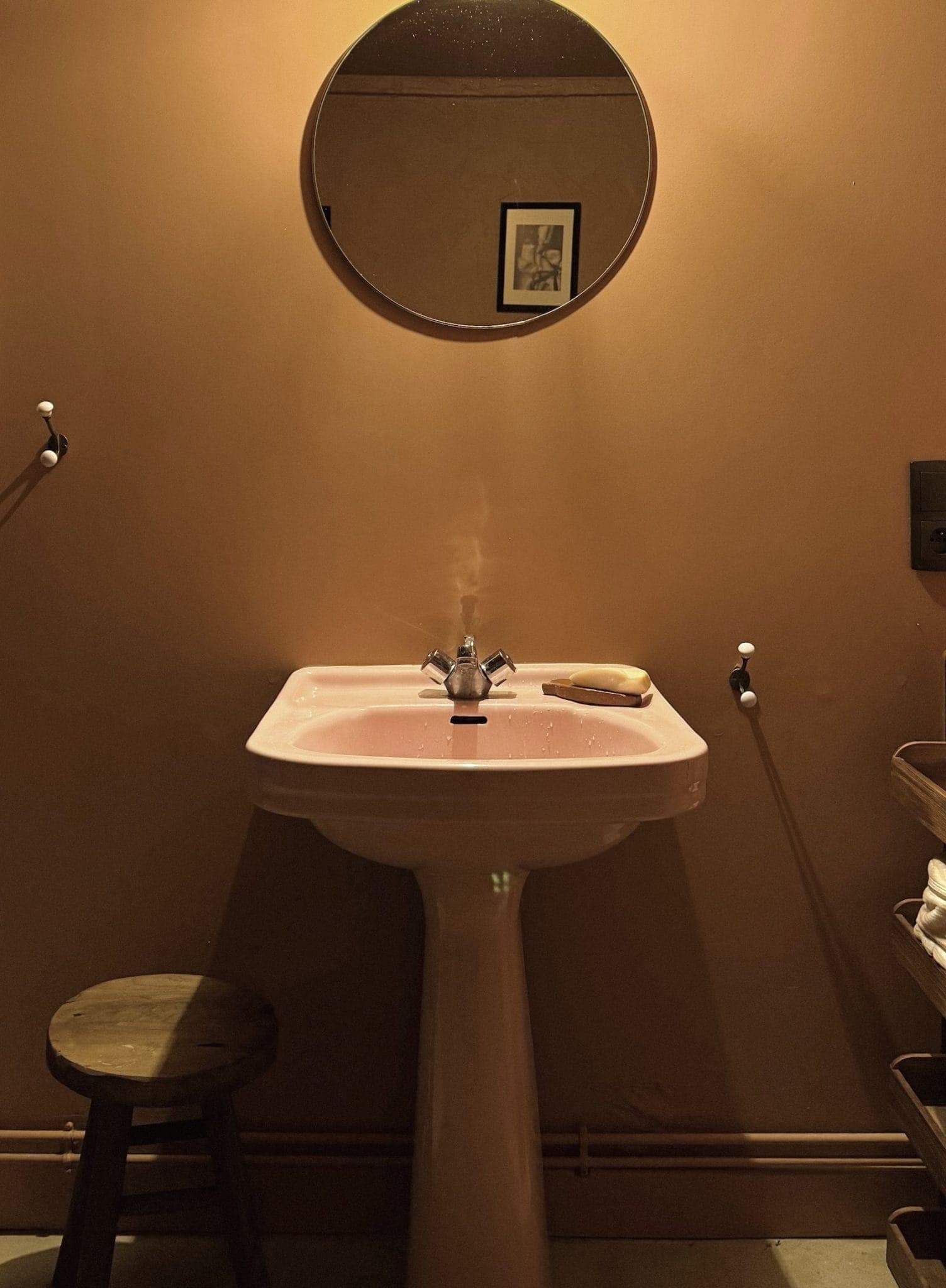 Une salle de bain aux murs orange chaleureux, avec un miroir rond au-dessus d'un lavabo sur pied rose. Un tabouret en bois se trouve à gauche et un porte-serviettes à droite. Un pain de savon repose sur le lavabo. Un tableau encadré se reflète dans le miroir.