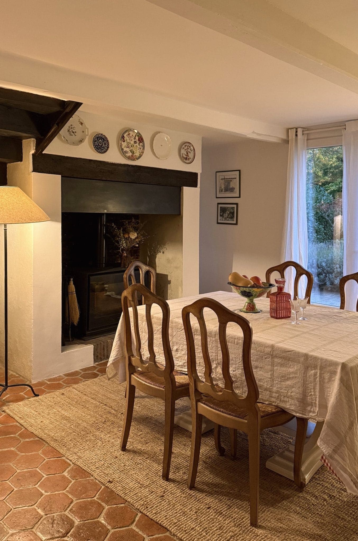 Une salle à manger chaleureuse avec une table recouverte d'une nappe blanche, entourée de chaises en bois. Une cheminée est décorée d'assiettes et d'œuvres d'art. Une lampe trône dans le coin et une grande fenêtre offre une vue sur la verdure extérieure.