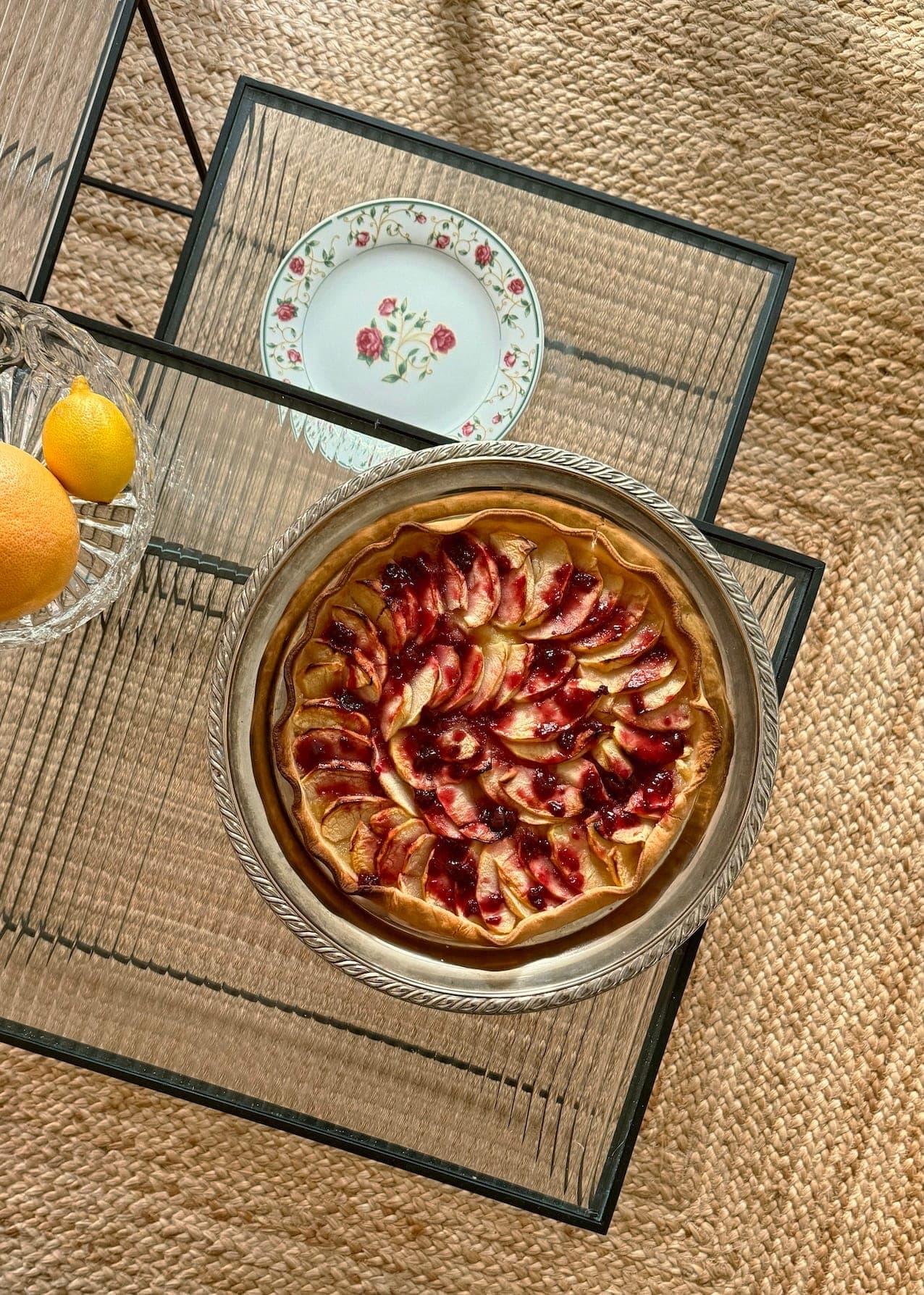 Une tarte aux pommes fraîchement cuite à la croûte dorée est posée sur une table basse en verre. À côté, on peut voir une assiette florale décorative et un bol contenant des citrons et une orange. La table repose sur un tapis tissé.