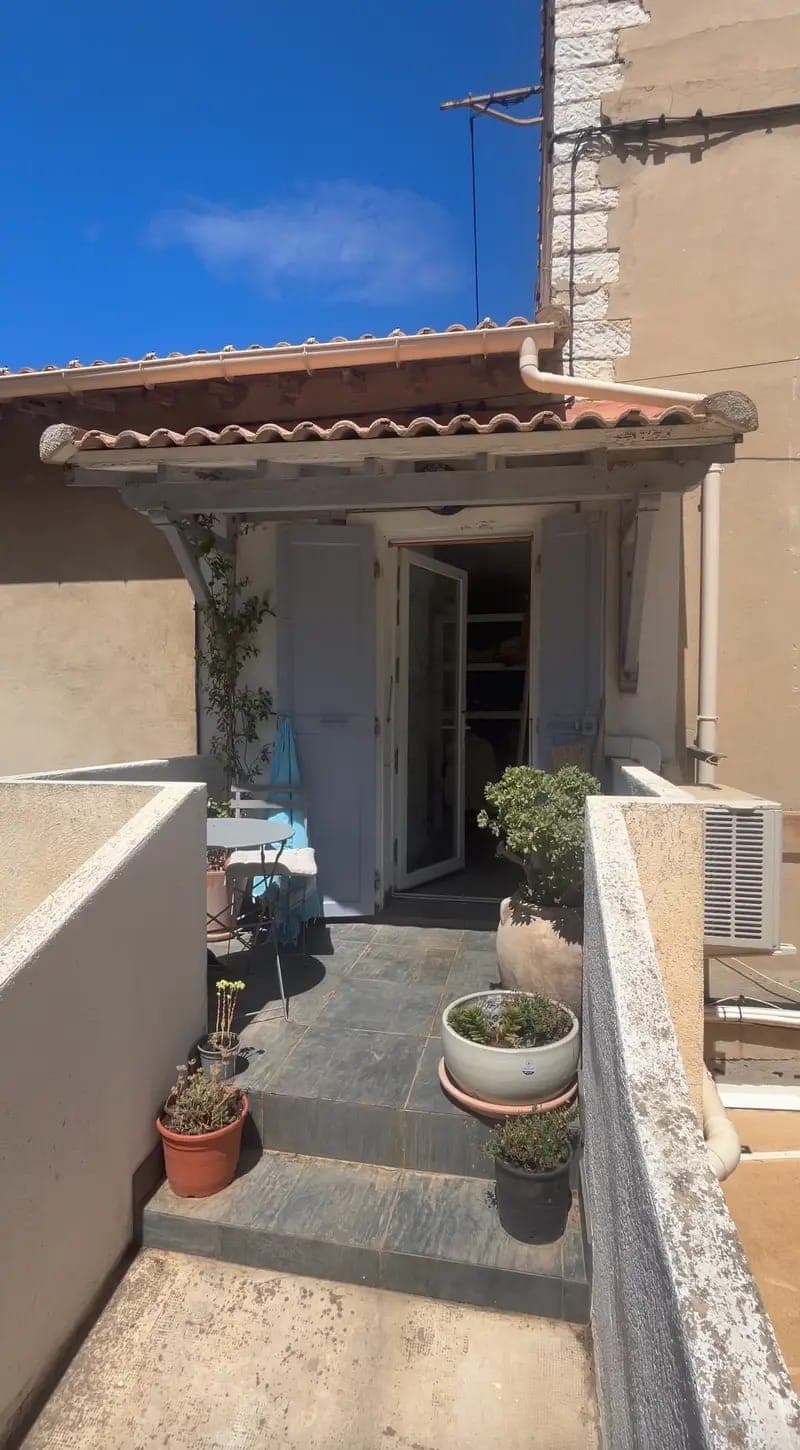 Une petite terrasse extérieure mène à une porte d'entrée avec des portes en bois ouvertes sous un toit en tuiles. Des plantes en pot bordent le chemin et une table avec une nappe bleue est placée près de l'entrée. Le bâtiment a des murs beiges et un climatiseur.