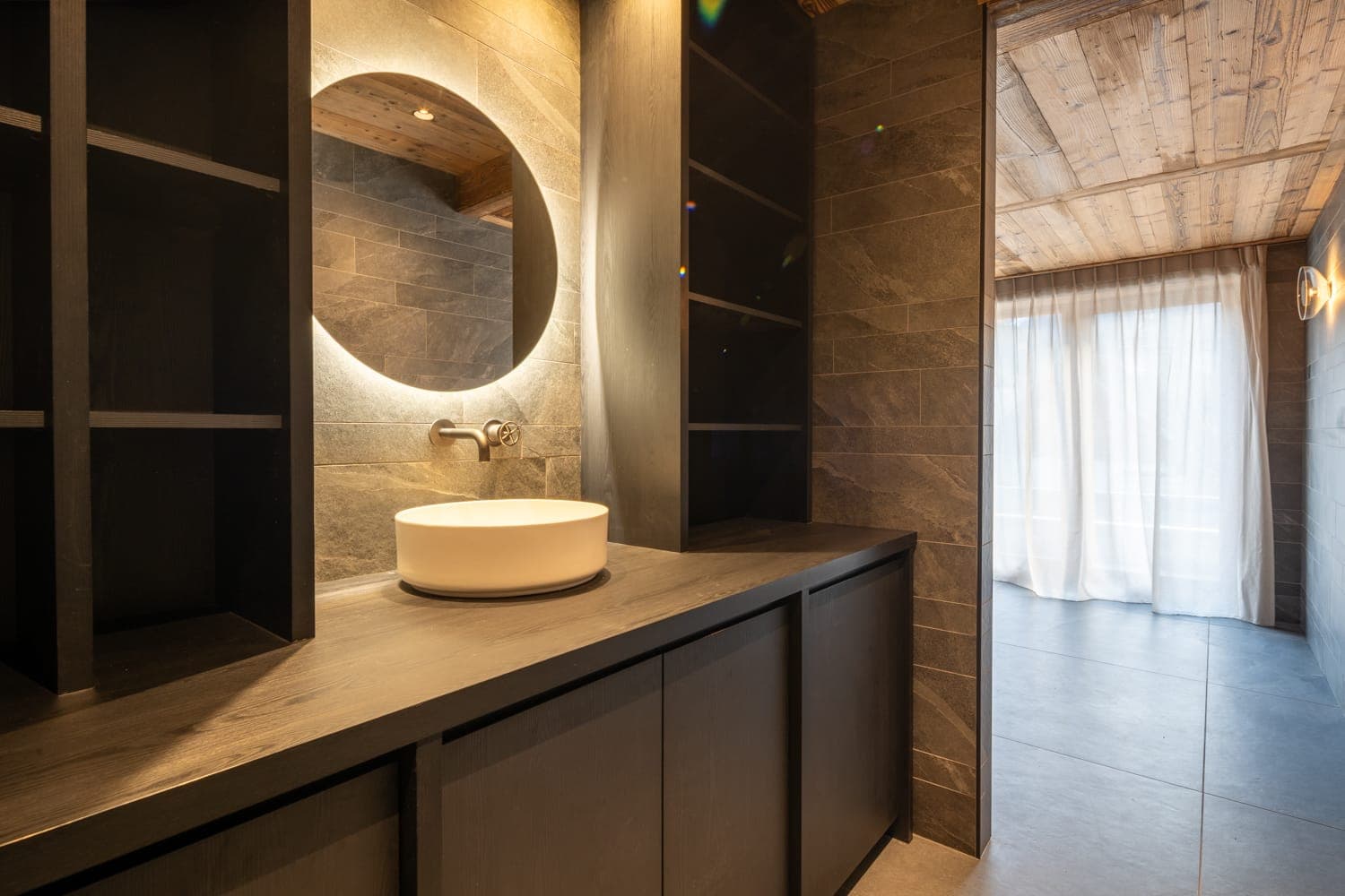 Salle de bains moderne avec murs en pierre et plafond en bois. Elle est dotée d'un miroir rond rétroéclairé au-dessus d'un lavabo blanc circulaire, posé sur un plan de travail foncé. La lumière naturelle filtre à travers des rideaux transparents recouvrant une grande fenêtre.