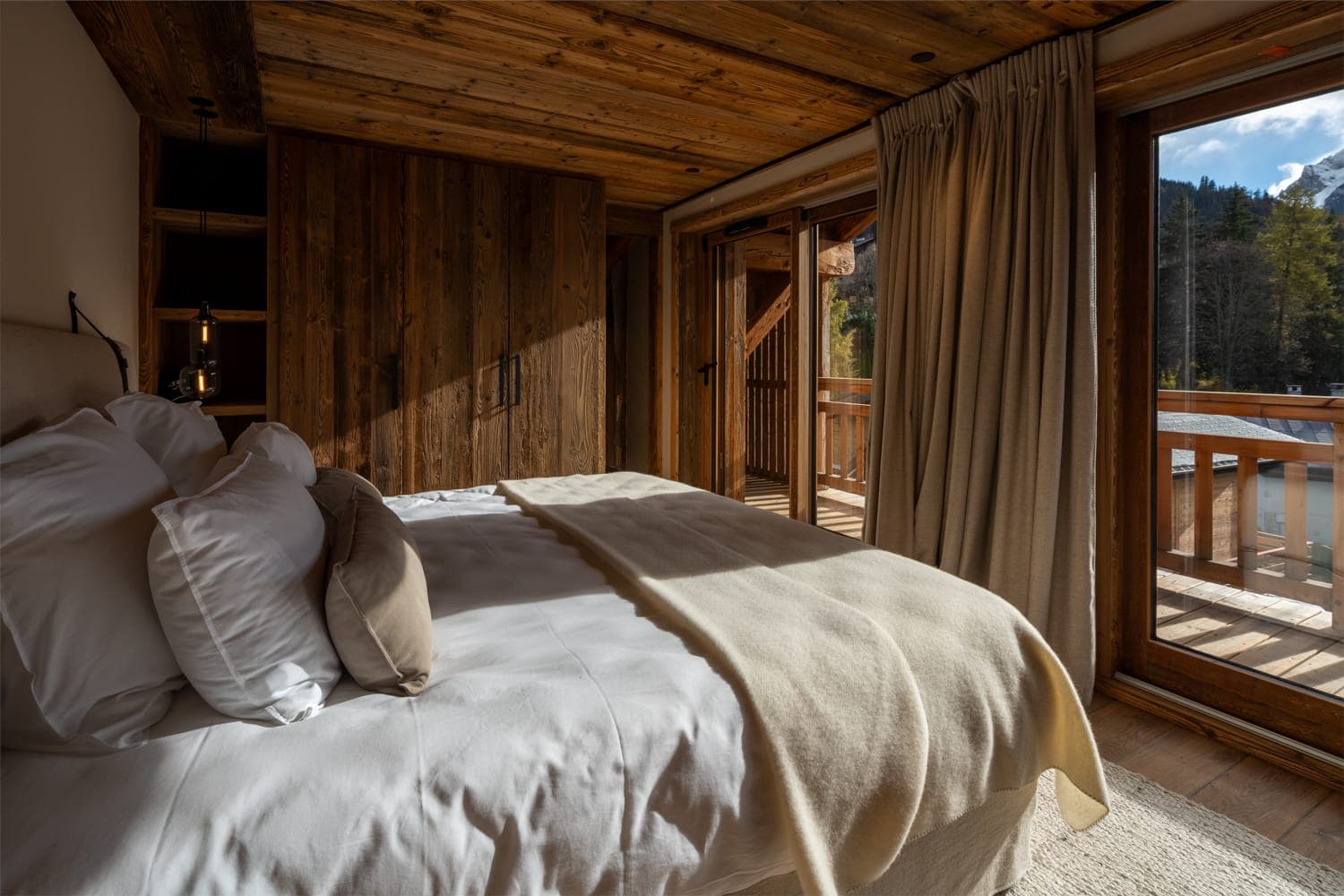 Une chambre confortable avec un plafond et des murs en bois rustique, dotée d'un lit soigneusement fait avec une literie beige et blanche. De grandes fenêtres avec des rideaux beiges offrent une vue sur un balcon en bois et des montagnes boisées en arrière-plan.