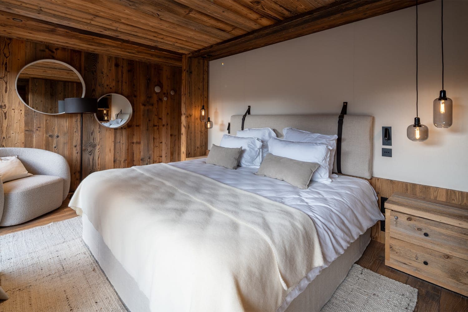 Une chambre confortable avec un intérieur en bois rustique. Elle comprend un grand lit avec une literie blanche et beige, une table de nuit en bois, deux miroirs ronds, des suspensions et un fauteuil beige. Les murs et le plafond sont en bois, ce qui ajoute de la chaleur à l'espace.