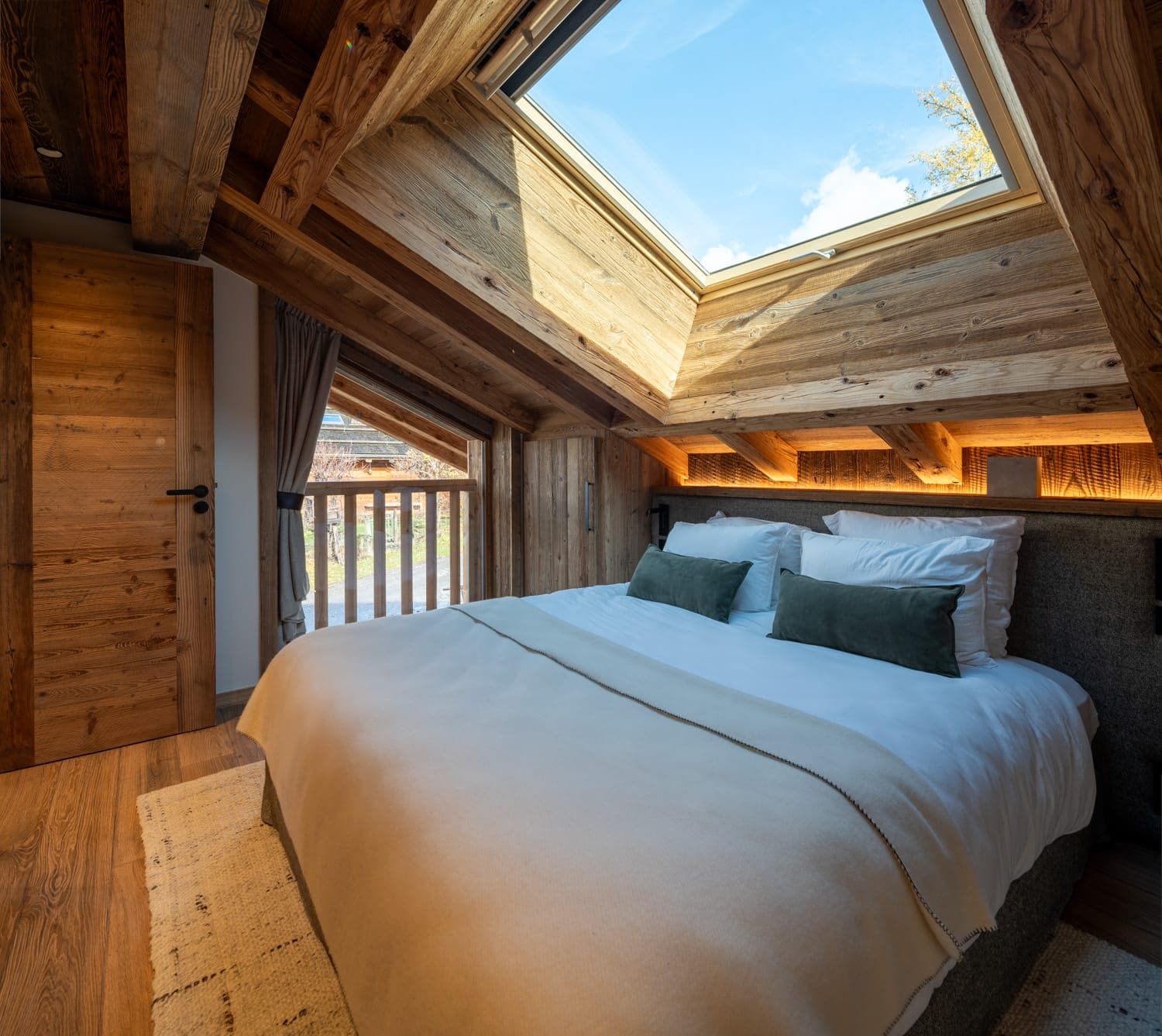 Une chambre mansardée confortable avec poutres en bois dispose d'un lit double habillé de literie blanche et verte. La pièce est éclairée par la lumière naturelle provenant d'une lucarne et d'une petite fenêtre. Une porte en bois et une balustrade de balcon sont visibles.