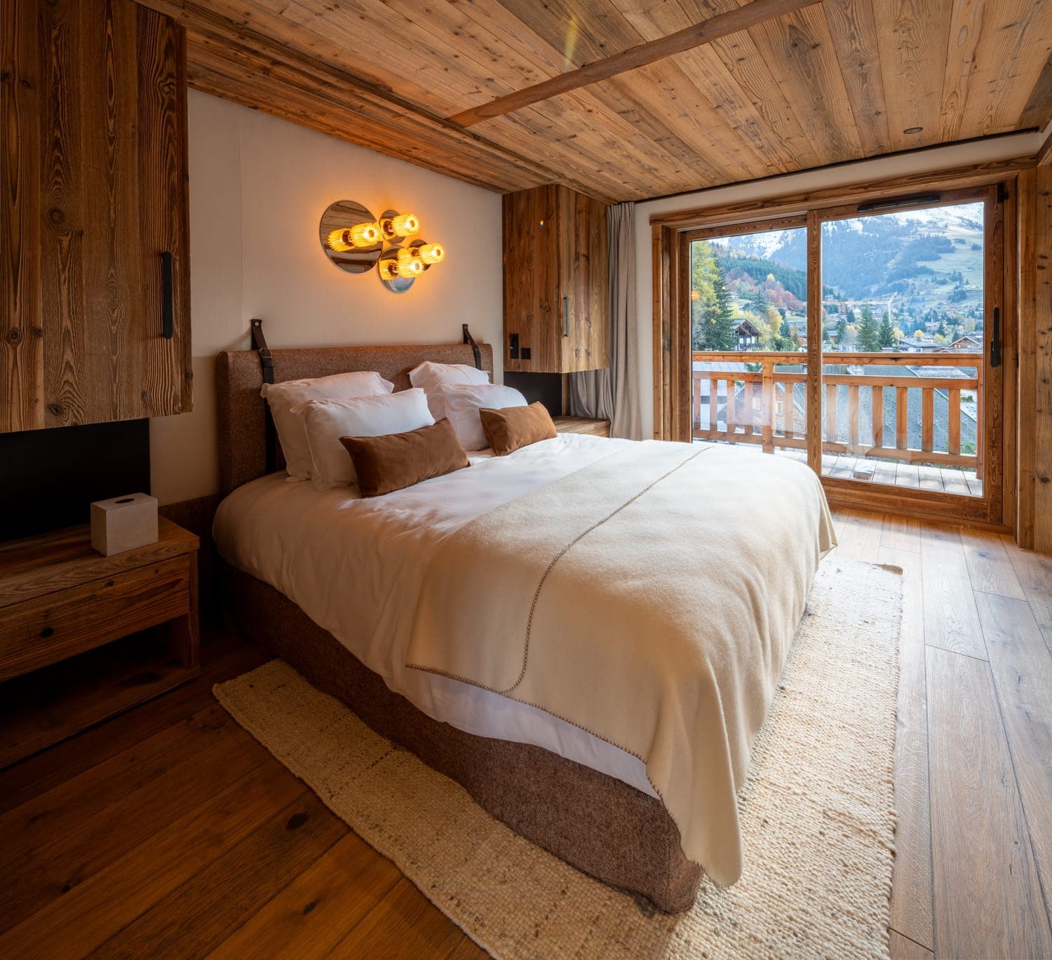 Chambre chaleureuse et rustique avec un grand lit, des murs et un plafond lambrissés de bois. Éclairage doux provenant d'un luminaire mural unique. Les portes vitrées s'ouvrent sur un balcon en bois avec vue sur un paysage montagneux pittoresque. La décoration aux tons terreux ajoute une atmosphère chaleureuse.