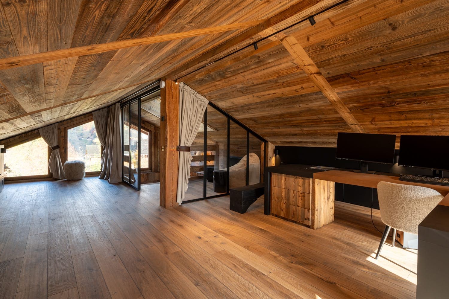 Un espace de travail mansardé confortable avec parquet et plafond en bois. Il dispose d'un grand bureau avec deux écrans et d'une chaise confortable. Des cloisons en verre séparent l'espace d'un petit salon avec une chaise moelleuse, donnant sur une vue panoramique sur l'extérieur.