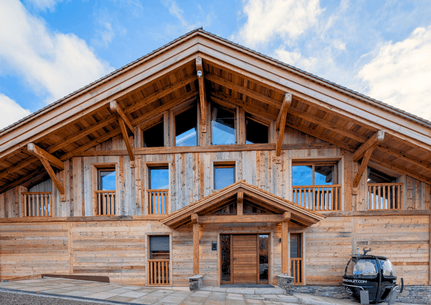 Un grand chalet rustique en bois avec un toit en pente raide et de nombreuses fenêtres sous un ciel bleu. Un téléphérique avec un téléski est visible à droite. Le décor évoque une zone montagneuse ou une station de ski.