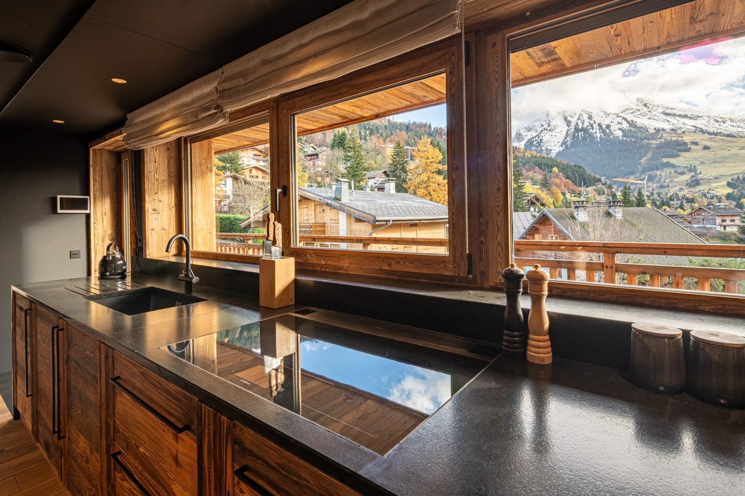 Une cuisine moderne avec un comptoir noir et des armoires en bois, comprenant un évier et une cuisinière. De grandes fenêtres révèlent une vue panoramique sur les montagnes enneigées, les maisons en bois et les arbres d'automne.