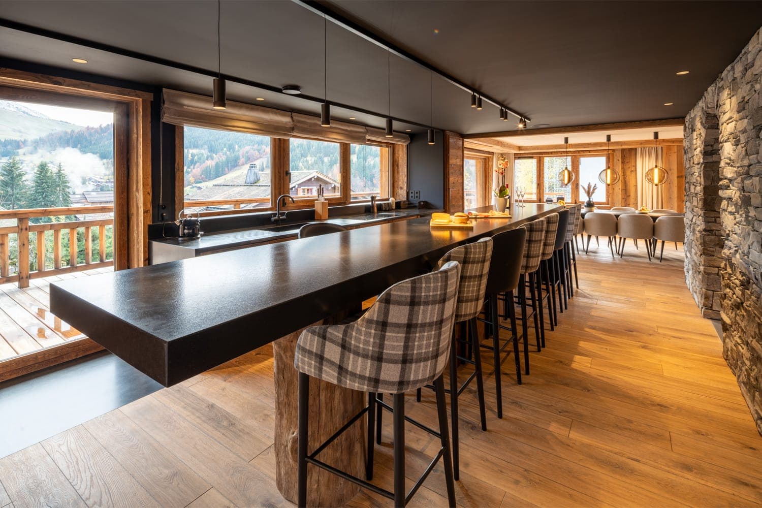 Cuisine et salle à manger modernes avec un long comptoir noir et des tabourets de bar à carreaux. De grandes fenêtres offrent une vue sur les montagnes et les cabanes en bois. L'espace présente des planchers en bois et des accents muraux en pierre, créant une ambiance chaleureuse et élégante.