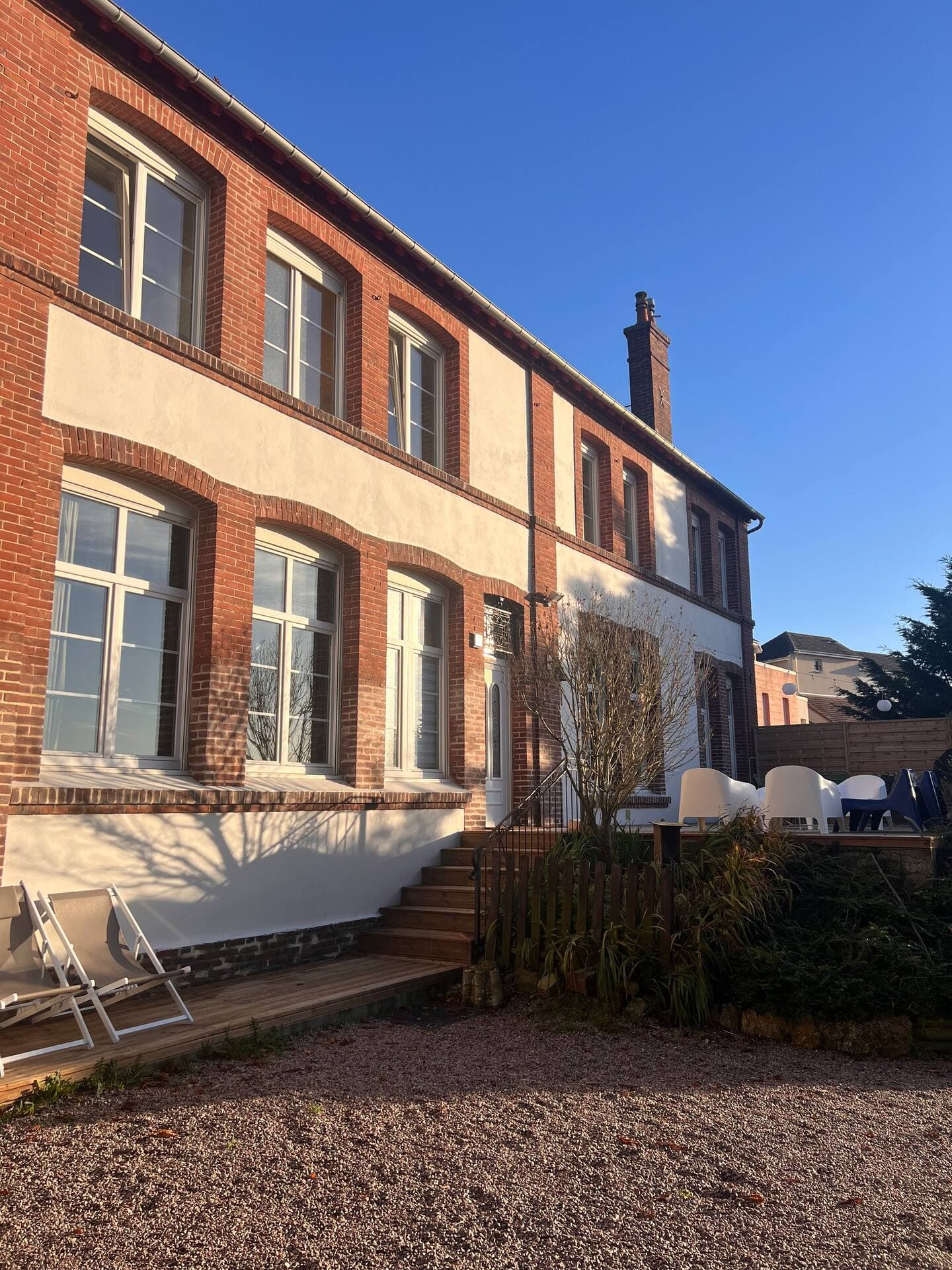 Un bâtiment de deux étages en briques rouges avec des touches de blanc, avec de grandes fenêtres. Un escalier mène à l'entrée et des chaises blanches sont disposées sur la terrasse. Le ciel est clair et bleu, ce qui indique une journée ensoleillée.