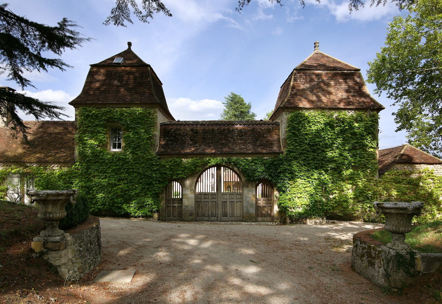 A rustic stone building with two towers, covered in green ivy, features a central arched entry with wooden gates. The structure is surrounded by trees and sits under a blue sky. Two planters are positioned symmetrically in the foreground.