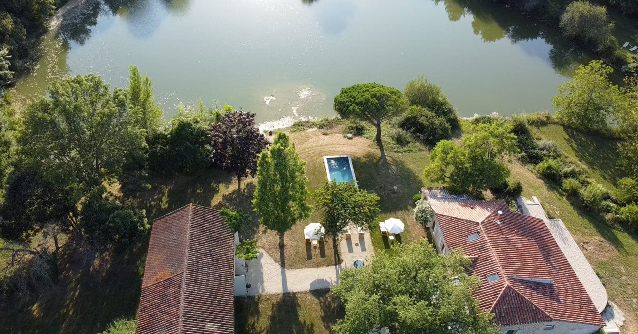 Vue aérienne d'une propriété sereine au bord d'un lac comprenant deux maisons aux toits rouges entourées d'arbres luxuriants. Une piscine rectangulaire est située entre les bâtiments, avec des chaises longues à proximité, le tout donnant sur un lac calme.