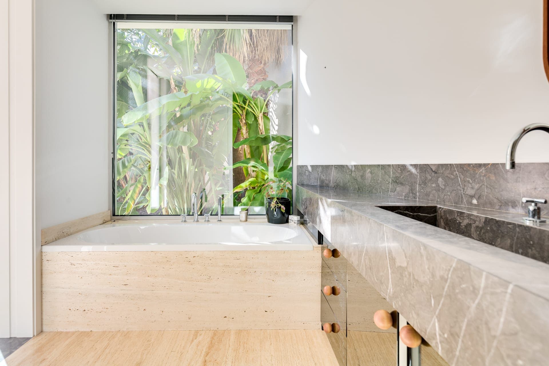 Salle de bain moderne avec une grande baignoire à côté d'une fenêtre laissant apparaître un feuillage vert luxuriant à l'extérieur. L'intérieur présente des comptoirs en marbre élégants, des accents de bois naturel et des éléments de design minimalistes.