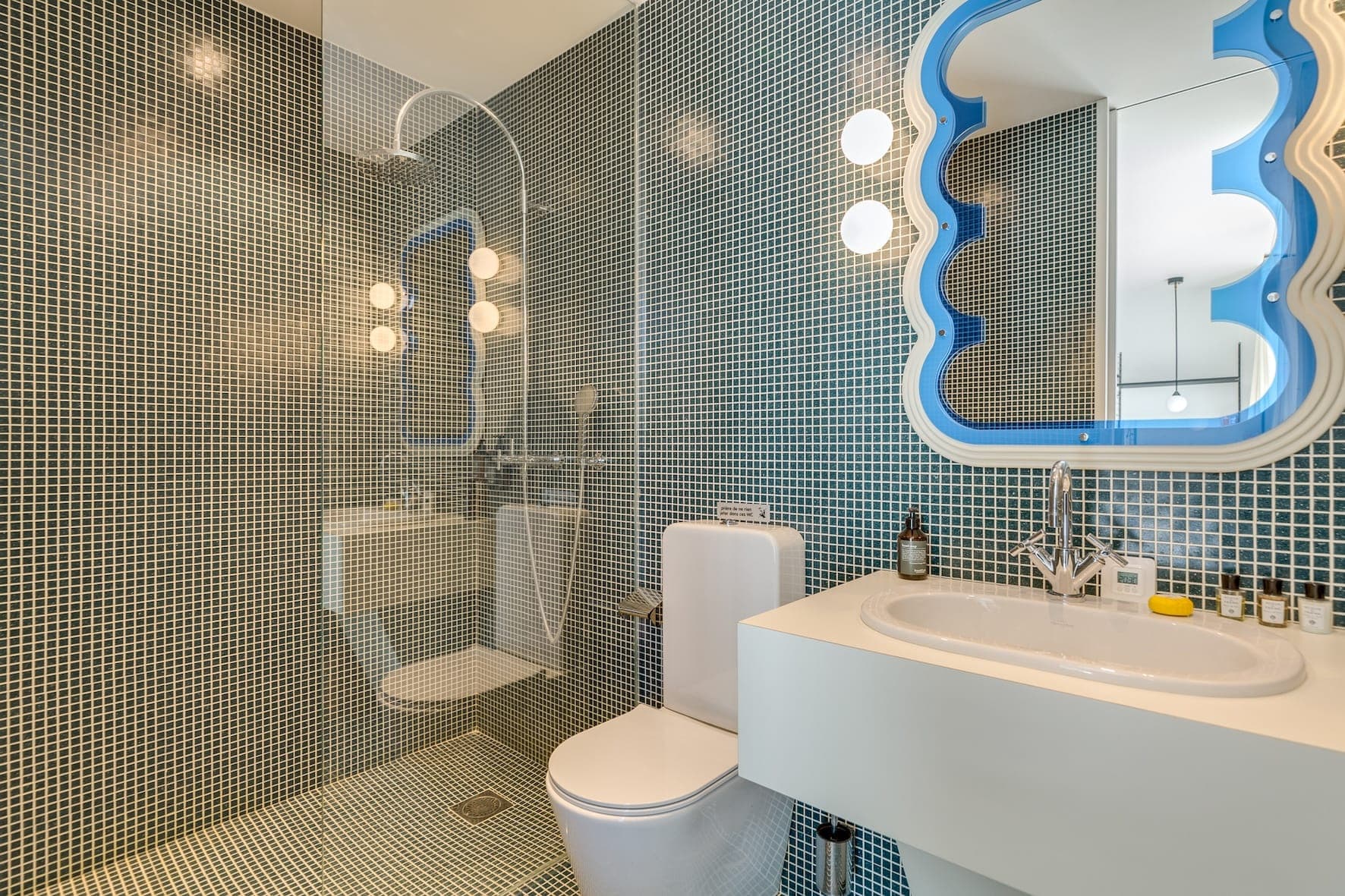 Une salle de bains moderne aux murs carrelés de bleu, avec une douche vitrée, des toilettes blanches et un lavabo blanc avec un élégant miroir ondulé à cadre bleu. L'espace est bien éclairé par des lampes à globe. Les articles de toilette sont soigneusement disposés sur le lavabo.