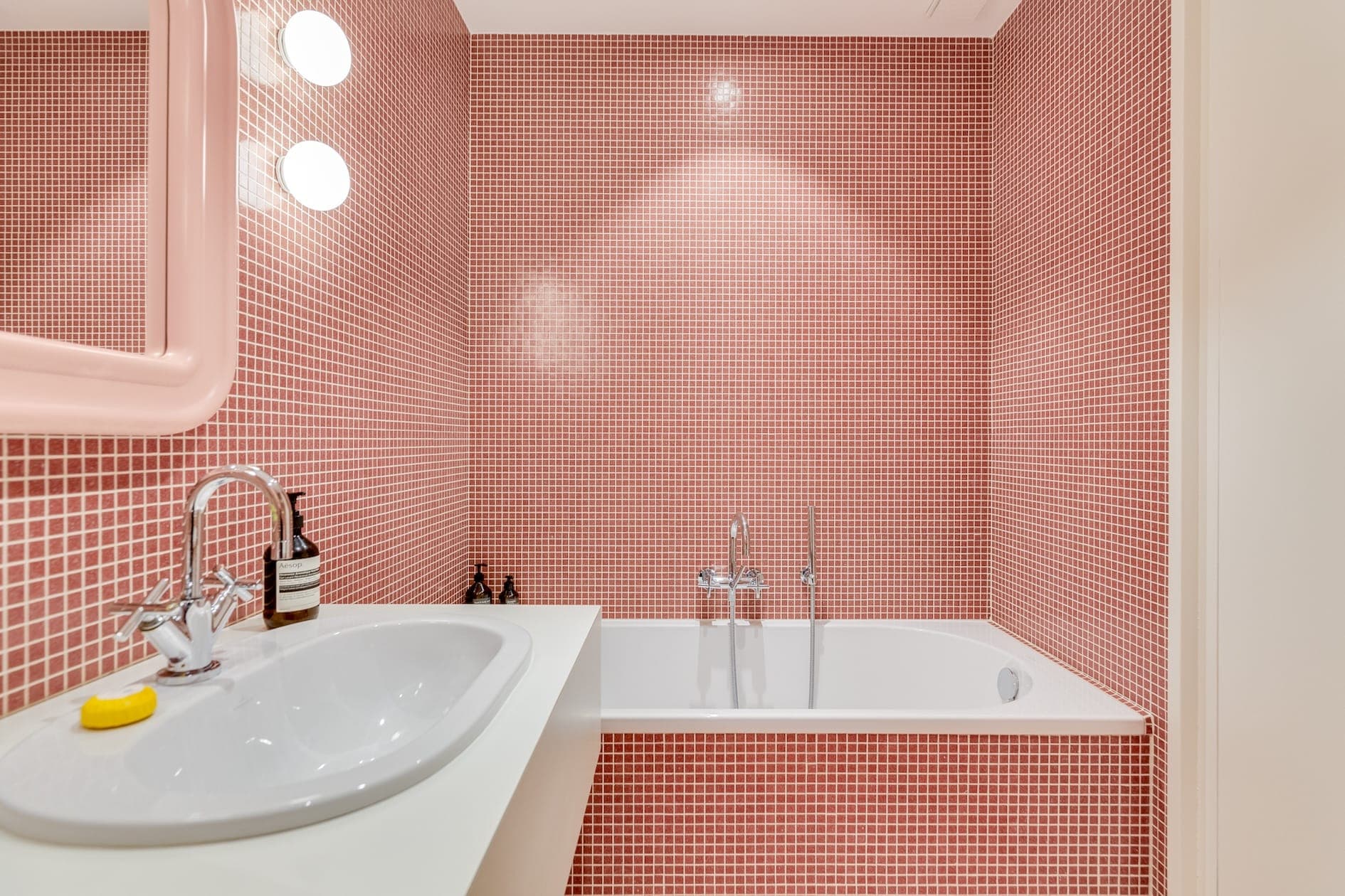 Une salle de bain avec des carreaux de mosaïque rose sur les murs, une baignoire blanche avec des robinets argentés, un miroir rectangulaire avec des lumières rondes au-dessus d'un lavabo et une bouteille de savon liquide sur le comptoir.