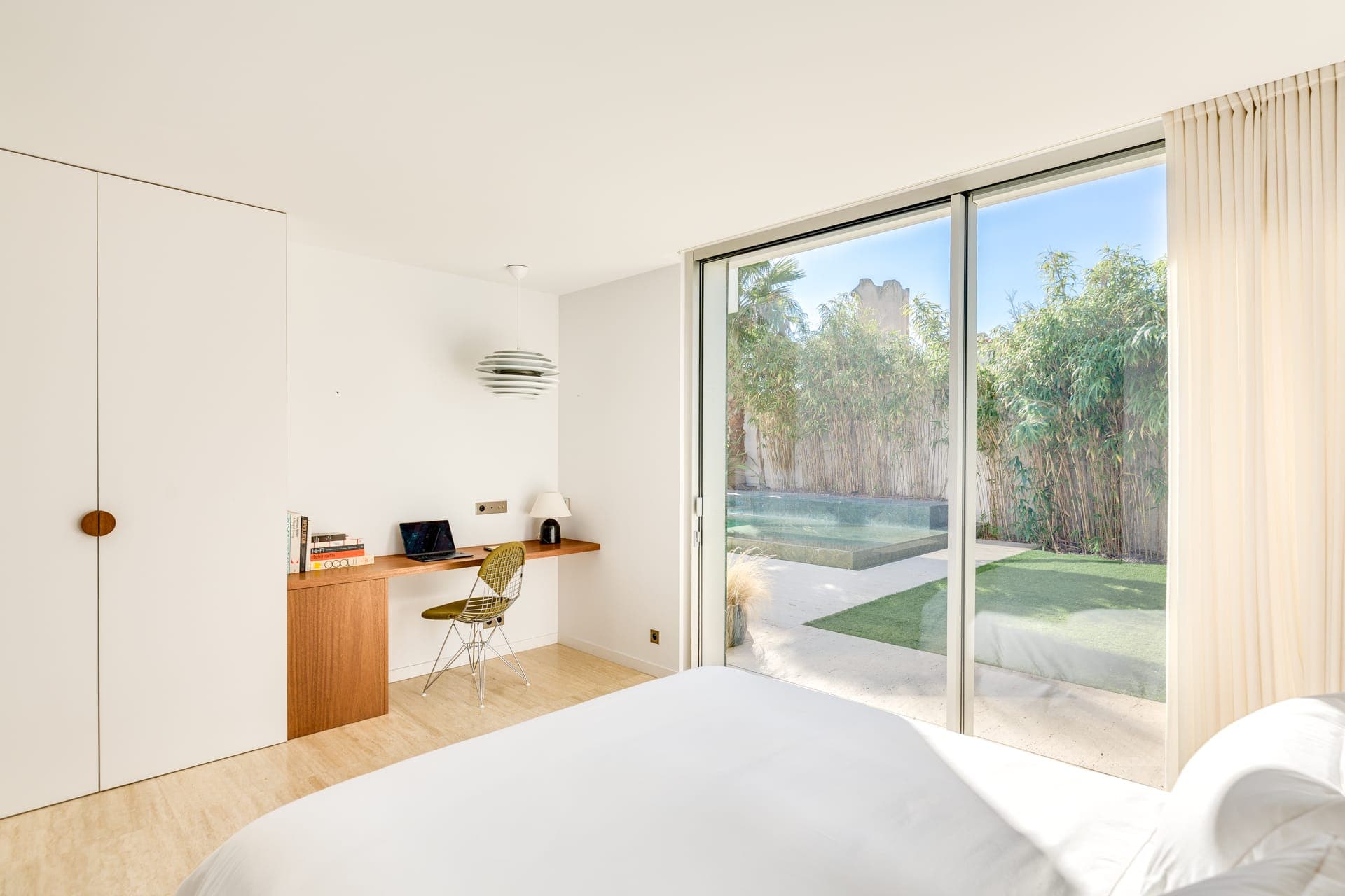 Chambre lumineuse avec un grand lit, des portes coulissantes en verre donnant sur un jardin avec une petite piscine et un coin bureau avec une chaise, une lampe et des livres. La chambre présente un design minimaliste avec des murs blancs et un parquet en bois clair.