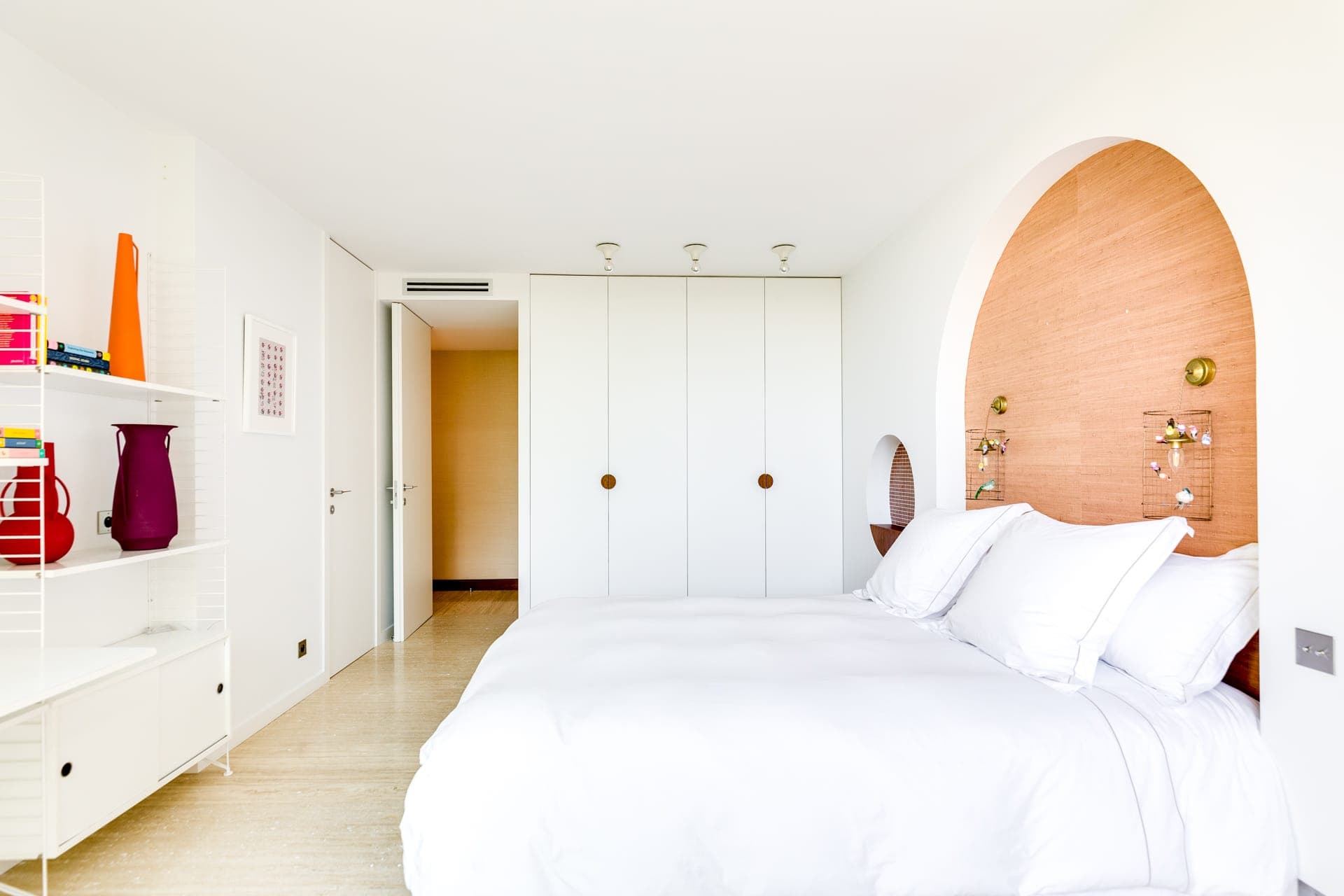 Une chambre moderne avec un grand lit blanc, des armoires encastrées blanches et un mur d'accent rond en bois derrière le lit. Les étagères à gauche contiennent des objets de décoration colorés et la pièce est lumineuse avec un design minimaliste.