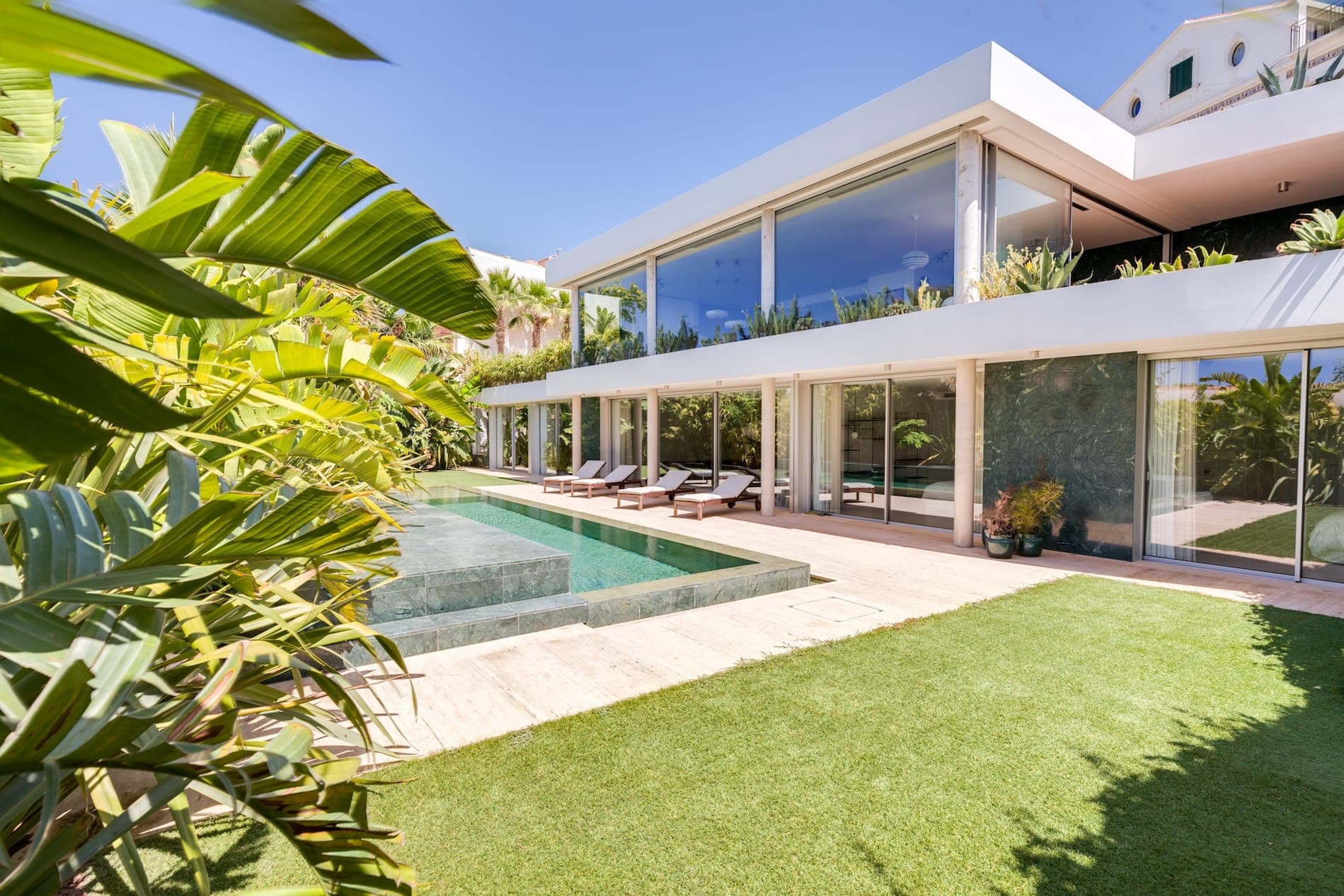 Une maison moderne à deux étages avec de grandes baies vitrées donnant sur une piscine rectangulaire entourée d'une végétation luxuriante et d'une pelouse soigneusement entretenue. La journée ensoleillée et lumineuse met en valeur le design architectural épuré.