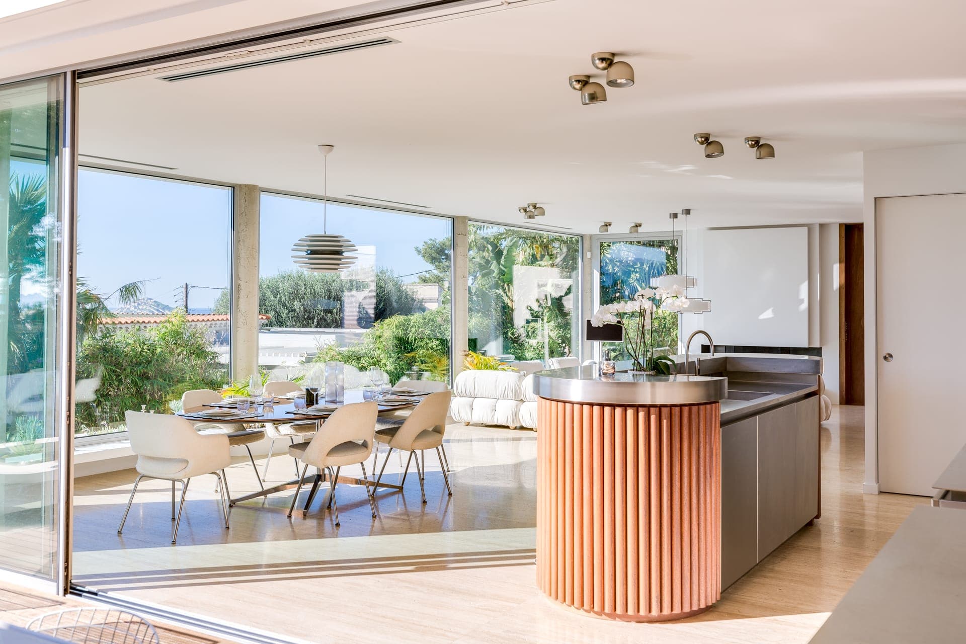 Spacieux espace de vie ouvert comprenant une cuisine moderne avec un îlot incurvé, une table à manger avec des chaises et un coin salon avec un canapé blanc. Les baies vitrées offrent une vue sur la verdure et une piscine extérieure. La lumière naturelle remplit la pièce.