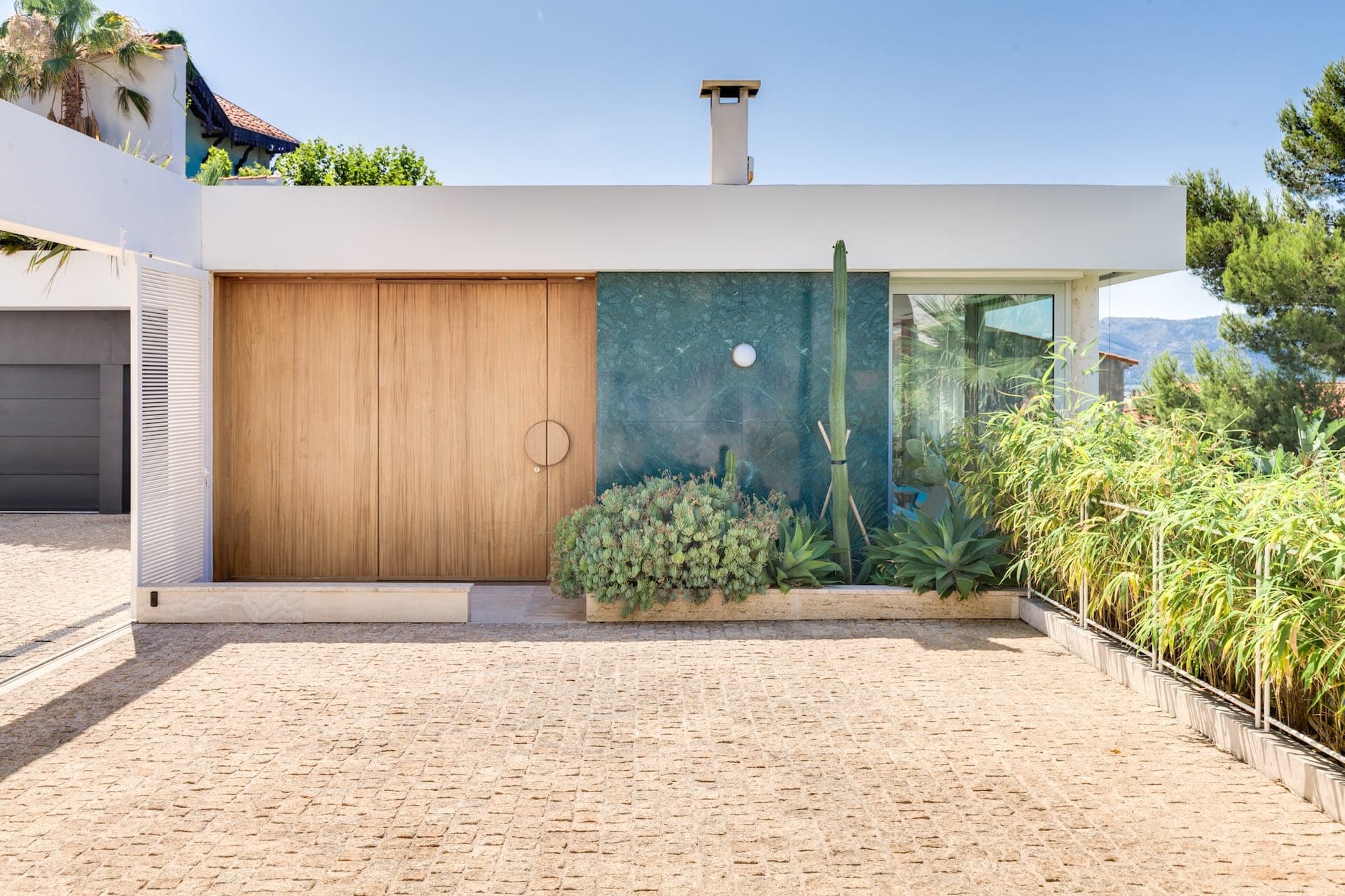 Entrée de maison moderne au design minimaliste, avec porte en bois, panneau mural végétalisé et plantes succulentes. Une allée pavée mène à l'entrée, entourée de verdure et d'arbres sous un ciel bleu clair.