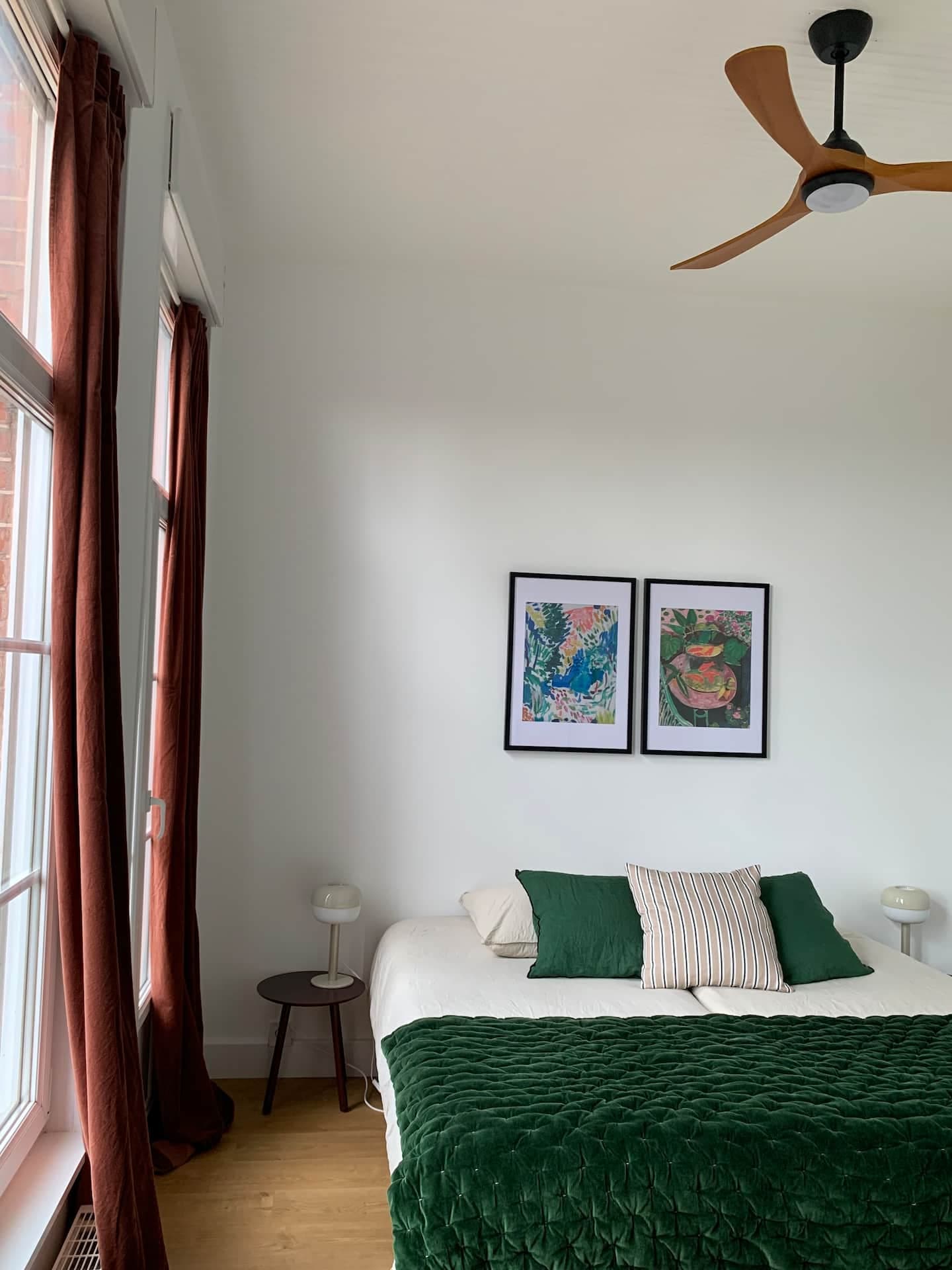 Une chambre avec un lit bien fait avec des draps et des oreillers verts. Deux œuvres d'art encadrées colorées sont suspendues au-dessus du lit. La chambre dispose de grandes fenêtres avec des rideaux marron et d'un ventilateur de plafond au-dessus. Une petite table d'appoint ronde avec une lampe se trouve à côté du lit.