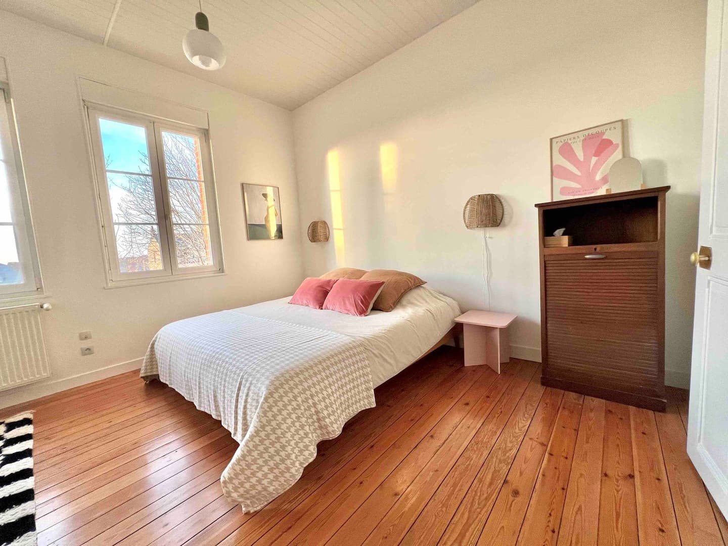 Une chambre confortable avec un lit double avec une couette blanche et beige, des coussins roses et une table de chevet rose. Une armoire en bois se trouve à côté. La chambre a du parquet, de grandes fenêtres et des œuvres d'art encadrées sur des murs blancs.