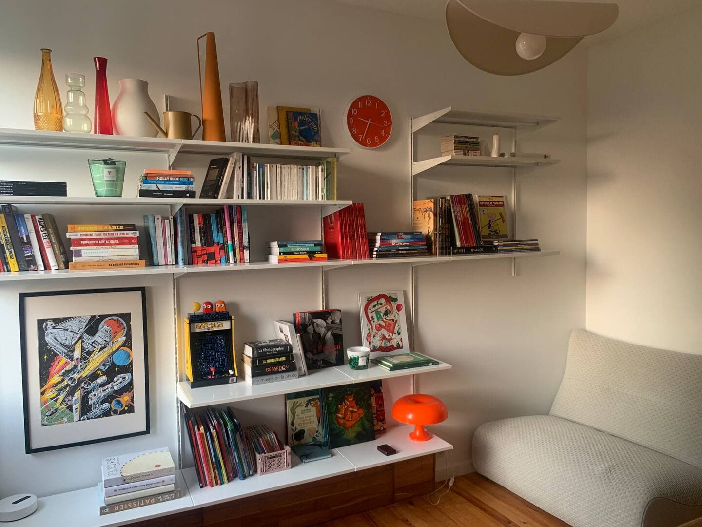 Une pièce confortable est dotée d'un mur blanc avec des étagères remplies de livres, de vases colorés et d'œuvres d'art. Une petite lampe de table orange est posée sur une étagère inférieure. Un luminaire moderne est suspendu au plafond et une chaise rembourrée de couleur claire se trouve dans le coin.