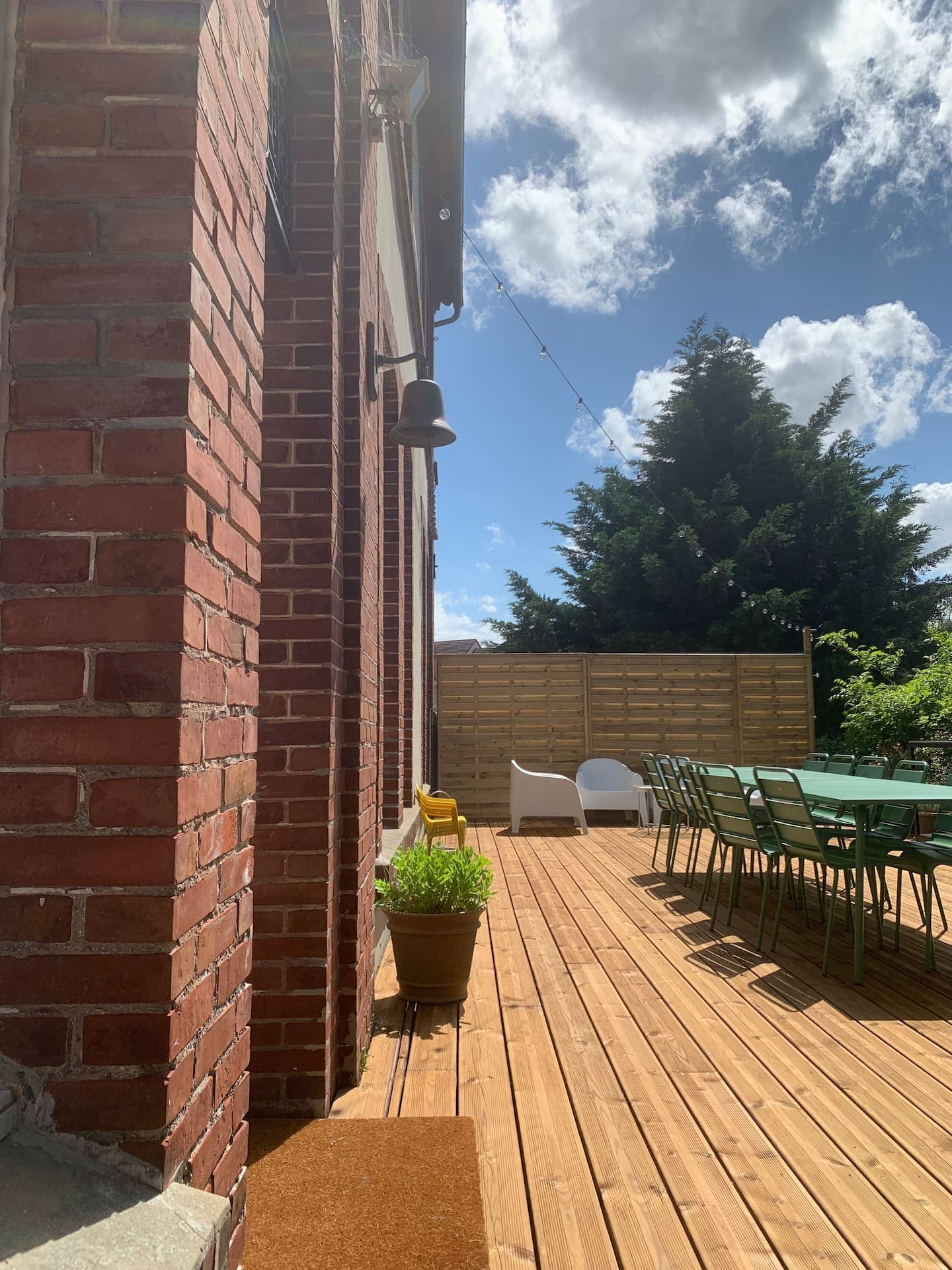 Une terrasse en bois ensoleillée à côté d'un bâtiment en briques rouges comporte une longue table verte avec des chaises. À l'arrière-plan, il y a deux chaises longues blanches et un grand arbre. Une petite plante en pot est posée près d'une applique murale. Le ciel est partiellement nuageux.