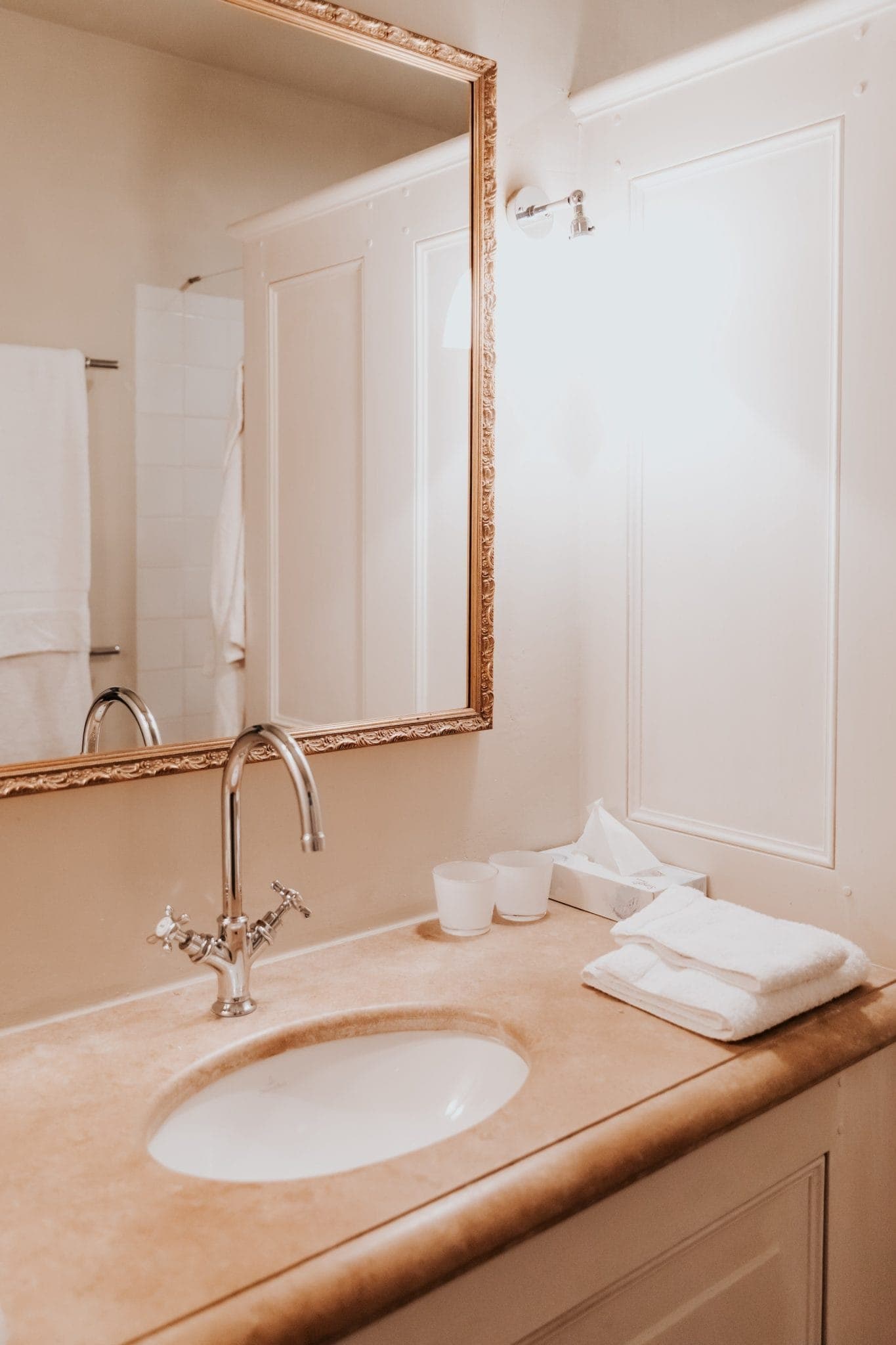 Salle de bain élégante avec plan de travail en marbre, lavabo encastré et miroir décoré. Un robinet en chrome poli se trouve au-dessus du lavabo. Une serviette, deux tasses blanches et une boîte à mouchoirs sont soigneusement disposées à côté. Un éclairage doux crée une ambiance chaleureuse.