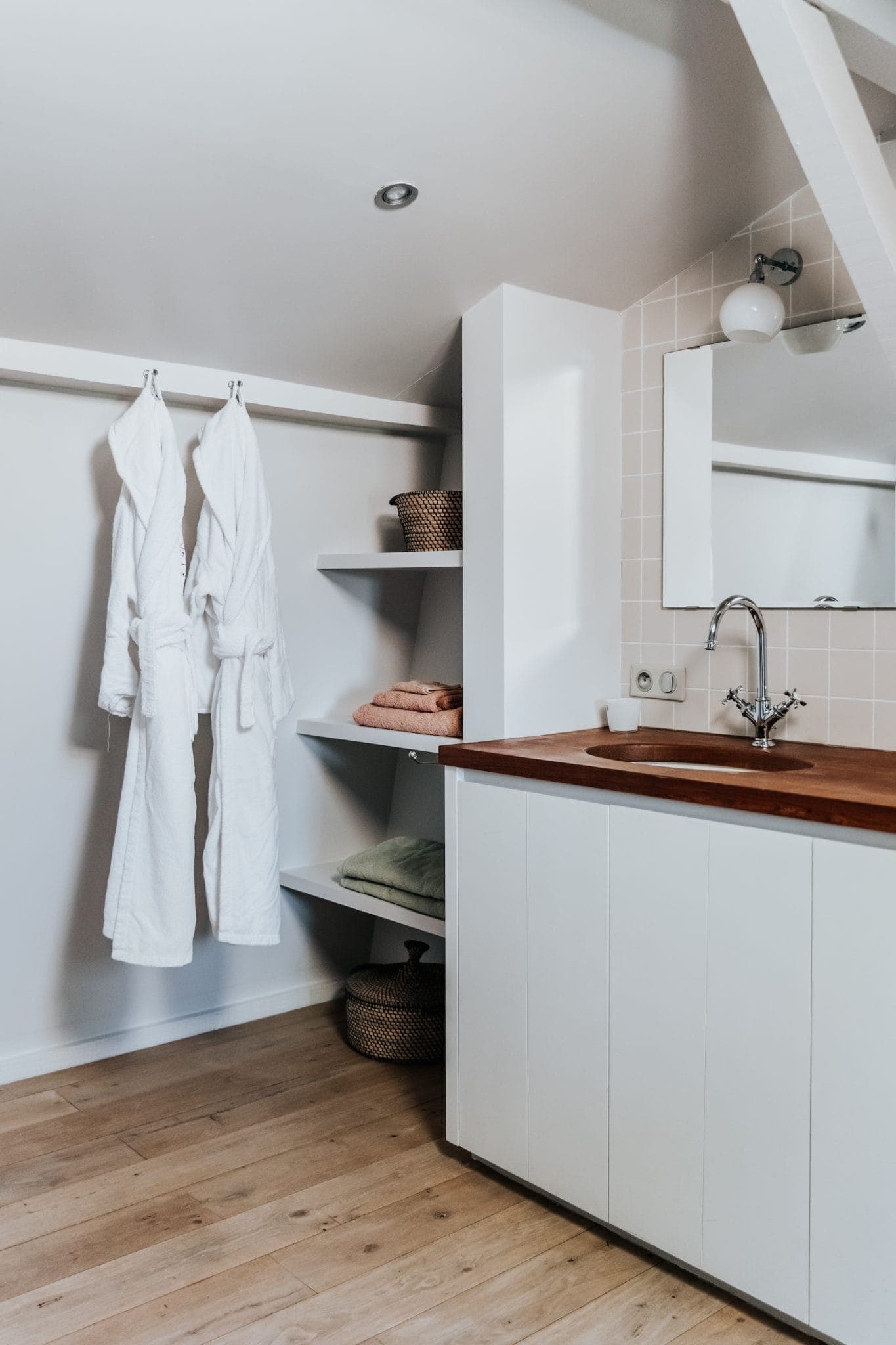 Une salle de bains minimaliste est dotée d'un parquet en bois clair, d'un plan de travail blanc avec une surface en bois, d'un lavabo mural, d'un grand miroir et d'un plafonnier. Deux peignoirs blancs sont suspendus à des patères à côté d'étagères ouvertes avec des paniers tressés et des serviettes pliées.