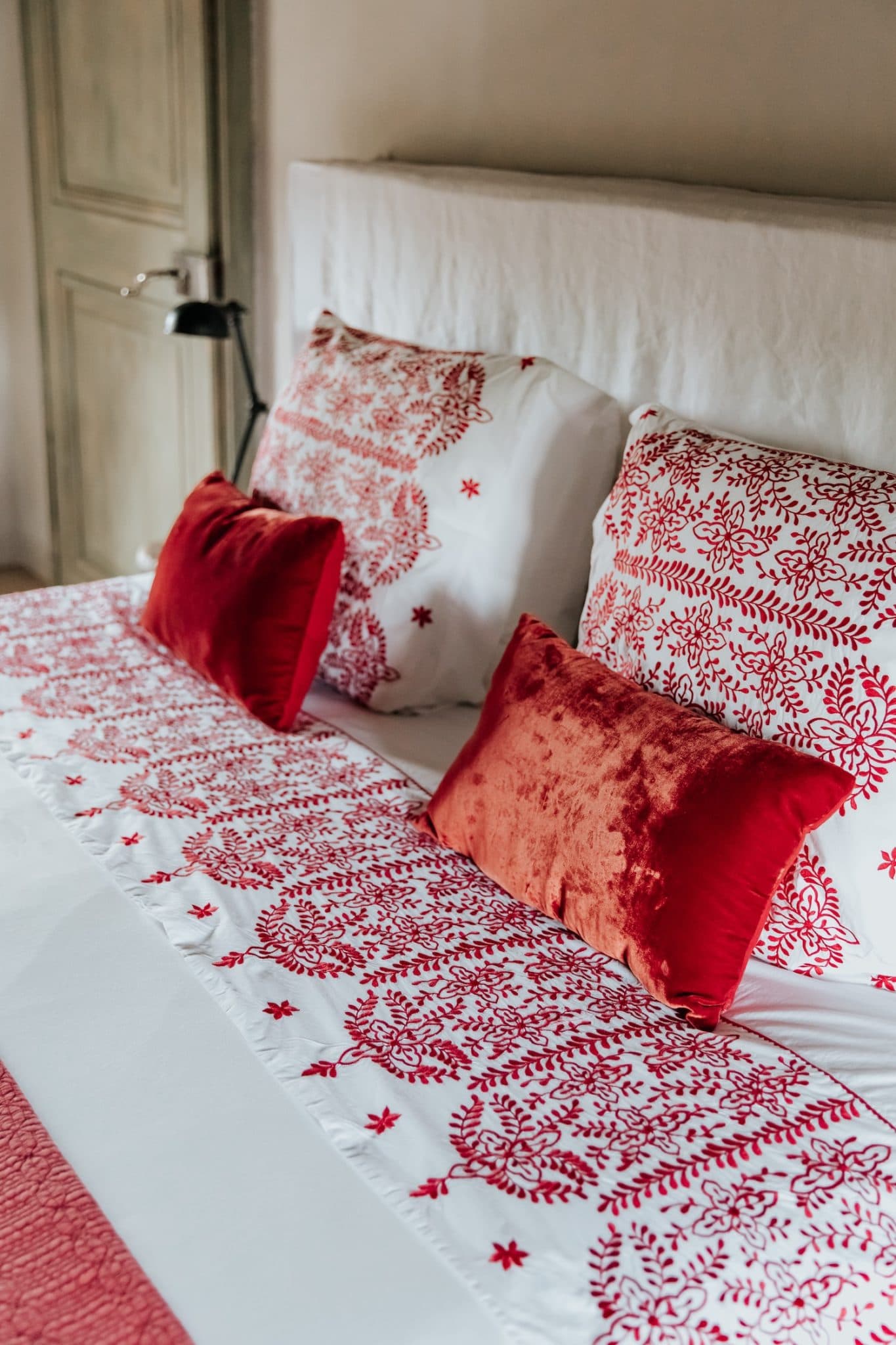 Un lit soigneusement confectionné avec des coussins décoratifs rouges et blancs et un chemin de lit assorti aux motifs complexes. Une lampe de lecture est visible à côté du lit et une porte en bois est partiellement visible à l'arrière-plan.