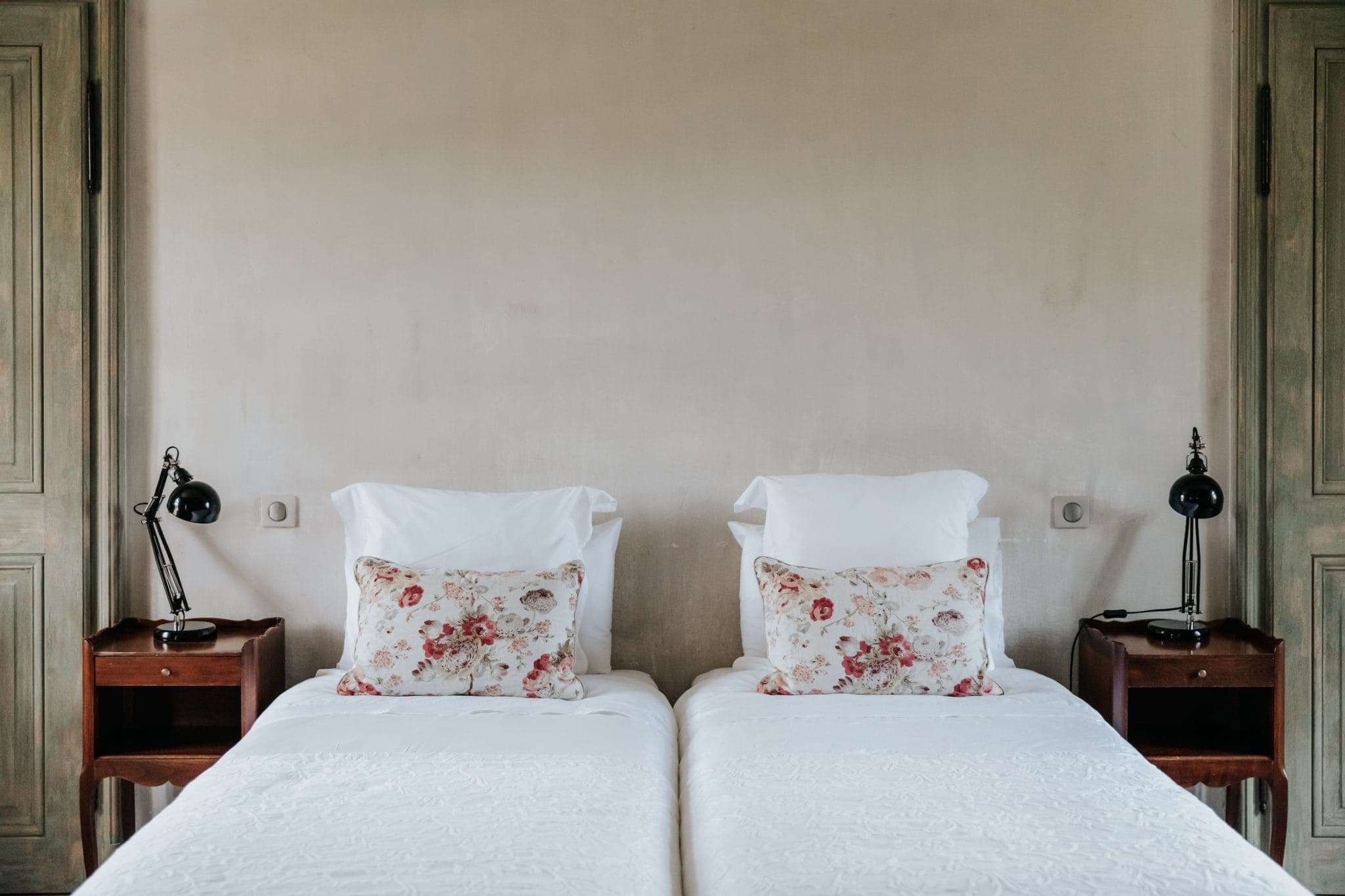 Une chambre confortable avec deux lits jumeaux recouverts de draps blancs et d'oreillers fleuris. Des tables de nuit en bois encadrent chaque lit, chacune surmontée d'une lampe de bureau noire. Les murs sont d'un beige discret et la décoration est simple et élégante.