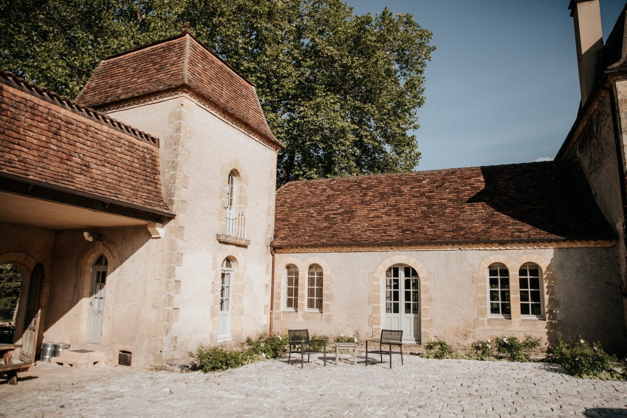 Une cour rustique en pierre entourée de bâtiments aux toits de tuiles rouges et aux fenêtres cintrées. Deux chaises sont placées au centre sur une surface pavée et des arbres verts luxuriants sont visibles en arrière-plan sous un ciel bleu clair.