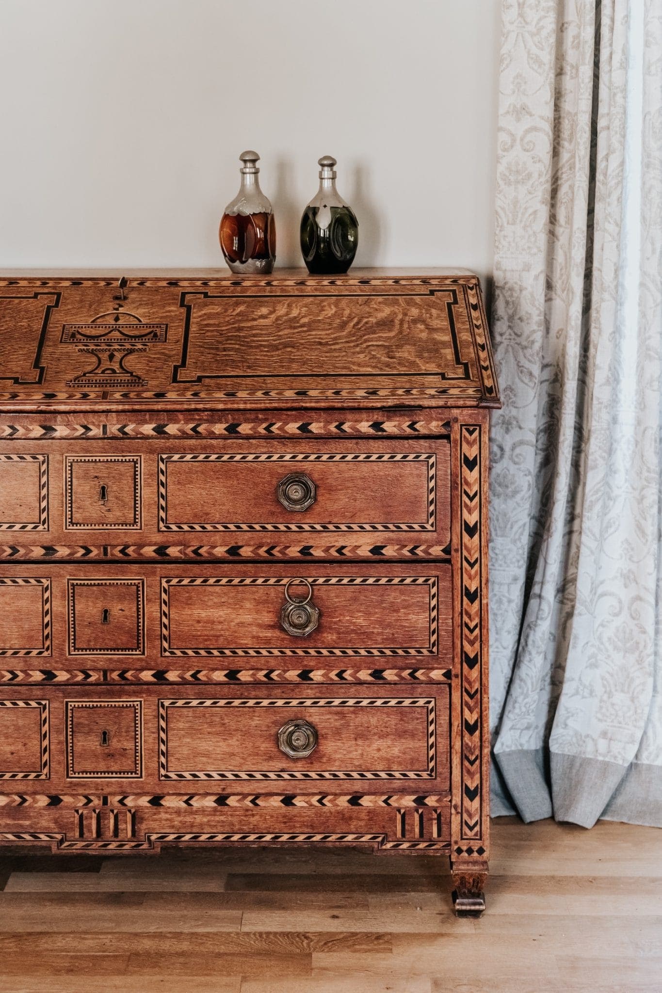 Une commode en bois ornée de sculptures complexes est adossée à un mur. Deux bouteilles décoratives reposent sur sa surface. Un rideau à motifs est partiellement visible sur le côté droit, avec un plancher en bois en dessous.