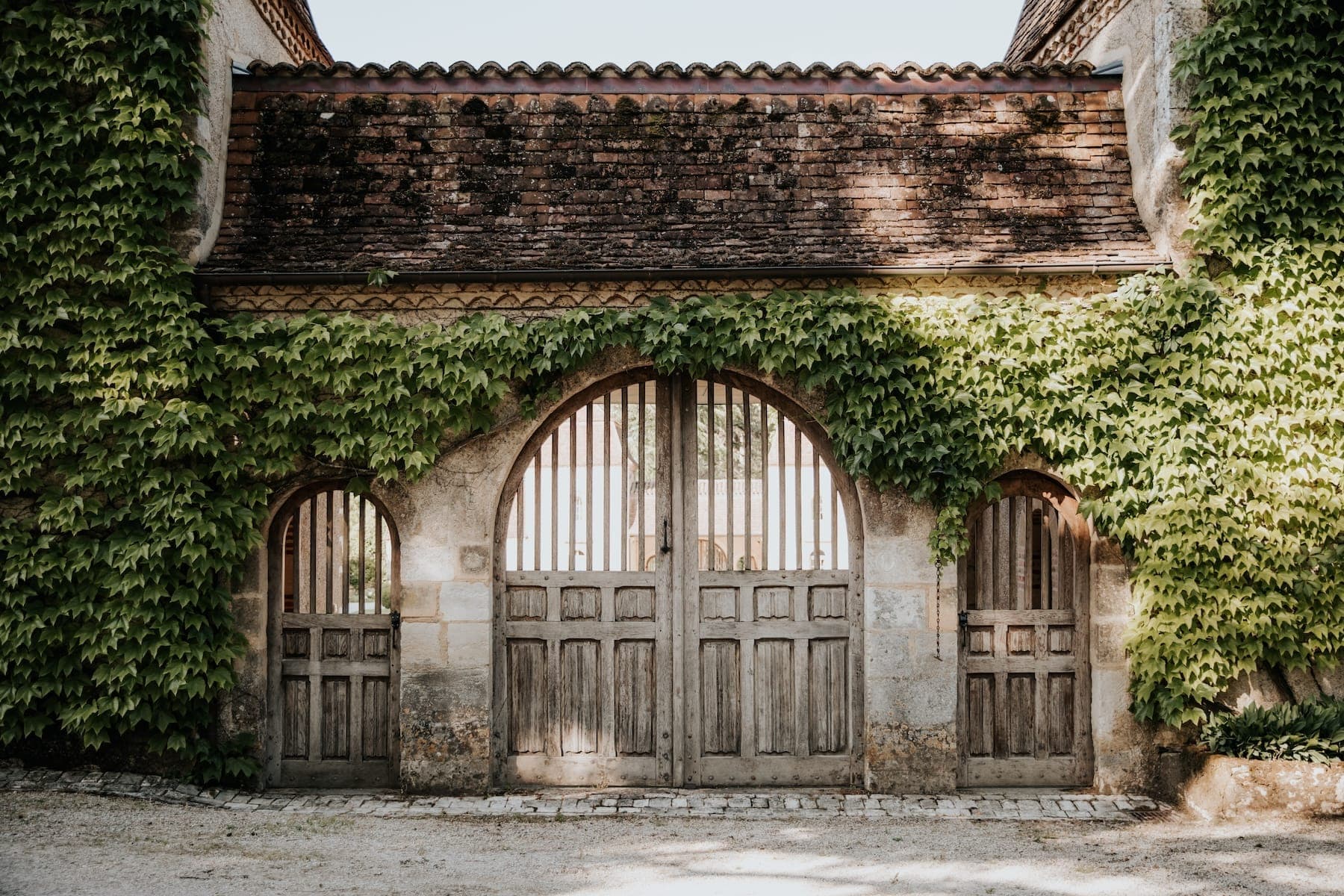 Un vieux bâtiment en pierre avec des portes en bois et des fenêtres cintrées, partiellement recouvert de lierre vert. Le bâtiment a une apparence rustique et patinée, et se trouve sous un ciel lumineux.