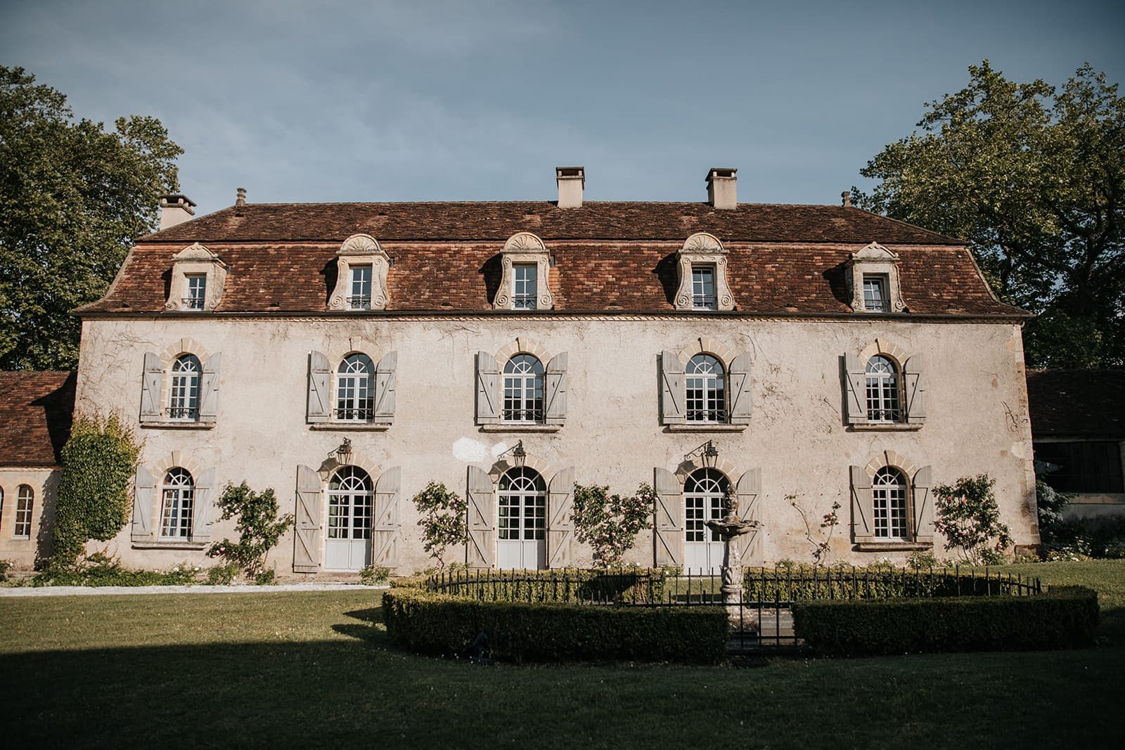 Un manoir historique en pierre de deux étages avec un toit en tuiles rouges, orné de multiples fenêtres cintrées. La façade présente un jardin bien entretenu avec une haie circulaire et une fontaine centrale, entouré d'une végétation luxuriante sous un ciel clair.